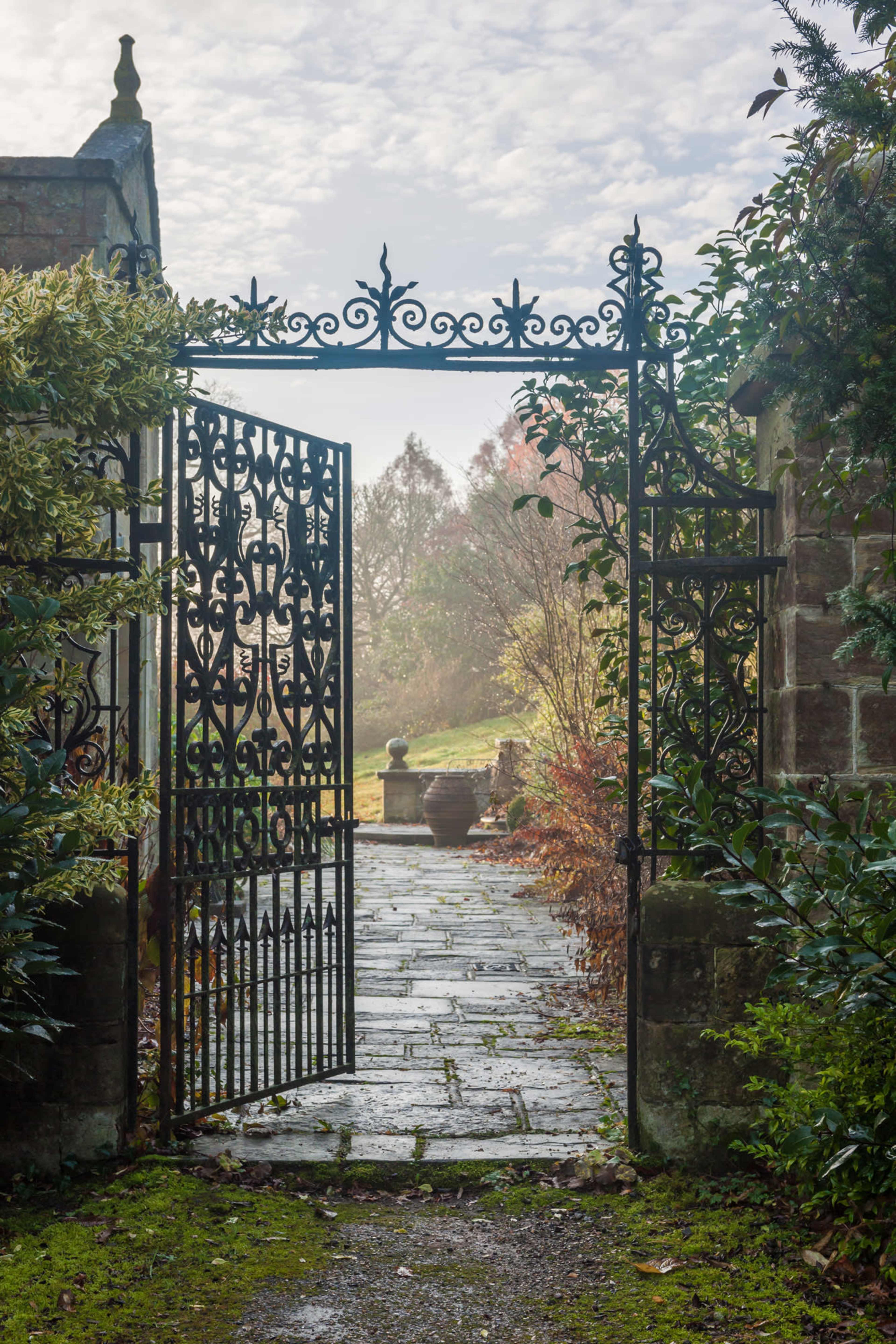 An ornate iron gate stands open, leading into a misty garden pathway lined with stone and surrounded by greenery.