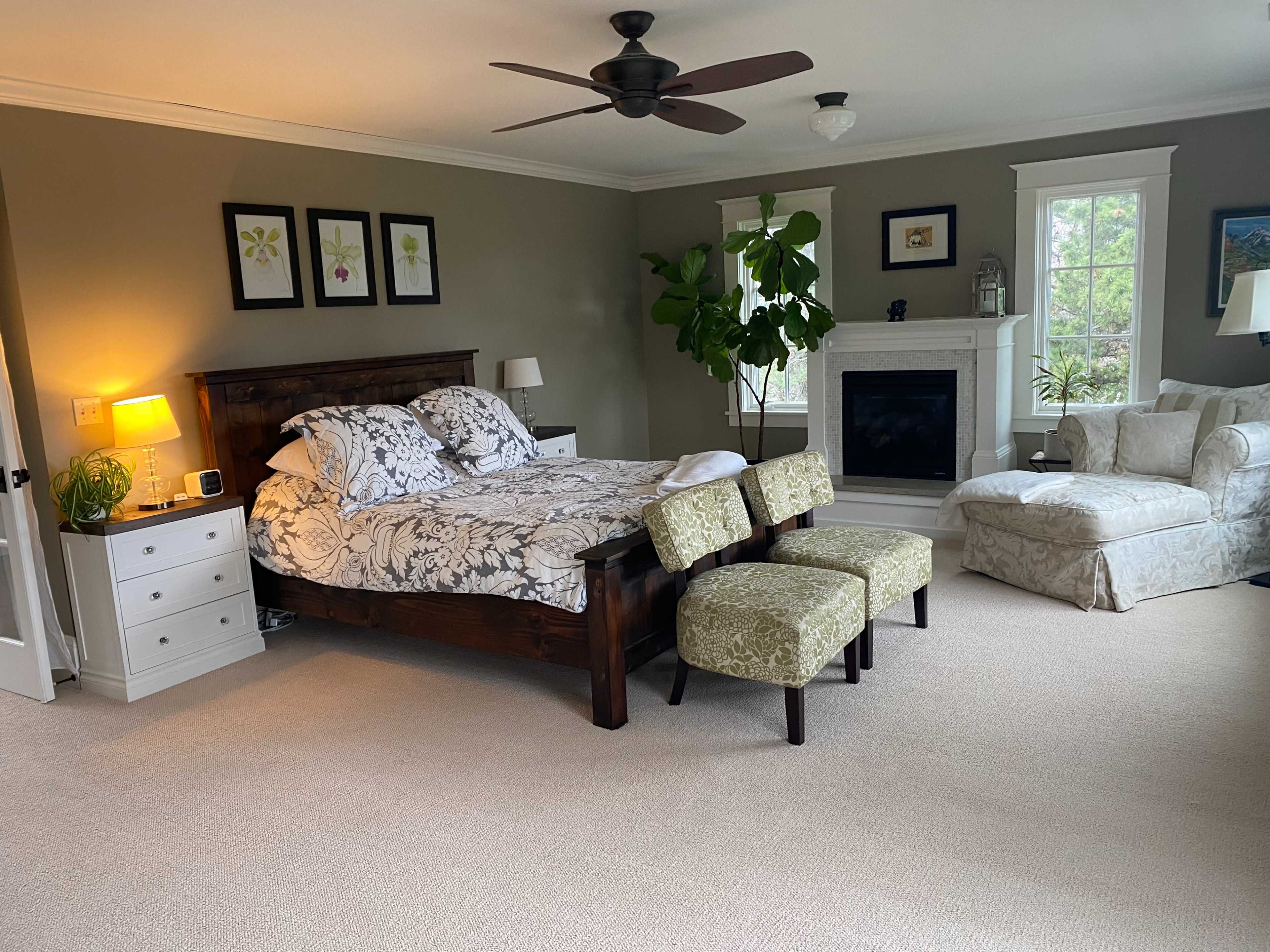 A spacious bedroom featuring a king-size bed with a floral duvet, two patterned stools at the foot, a white armchair, a fireplace, and large windows.