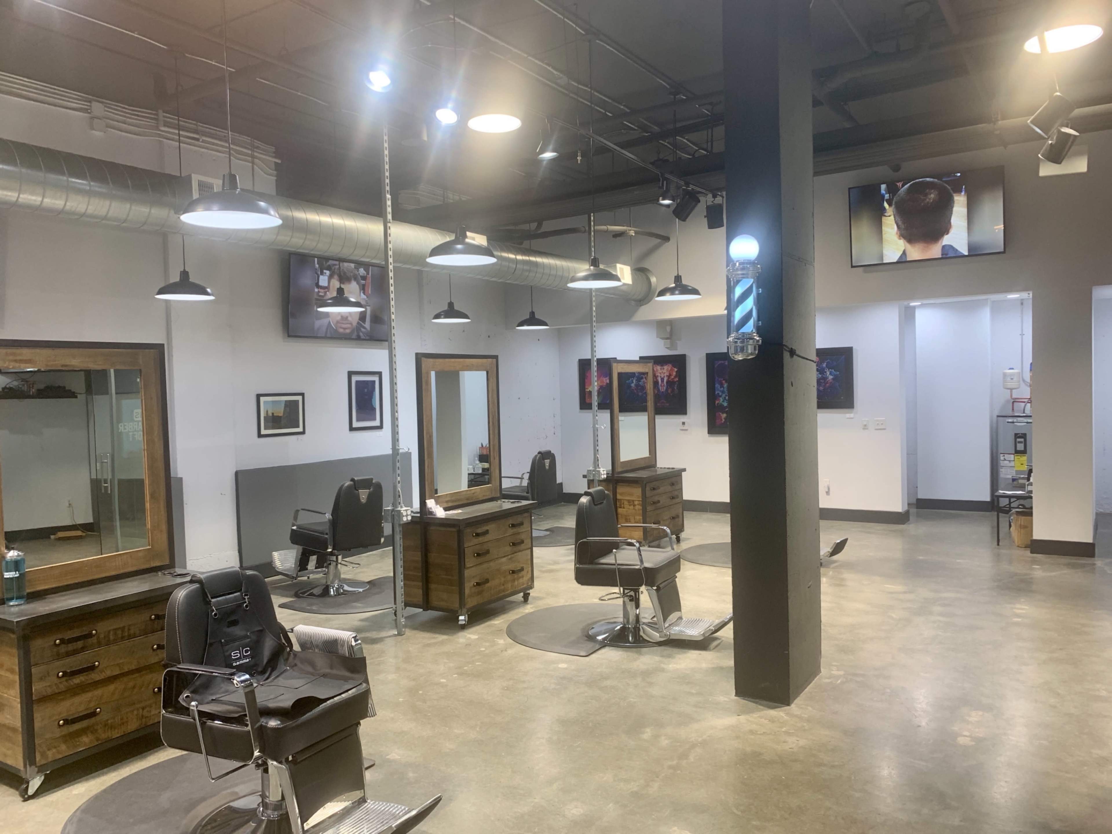 The image shows a modern barbershop interior with multiple styling stations, mirrors, and flat-screen TVs on the walls.