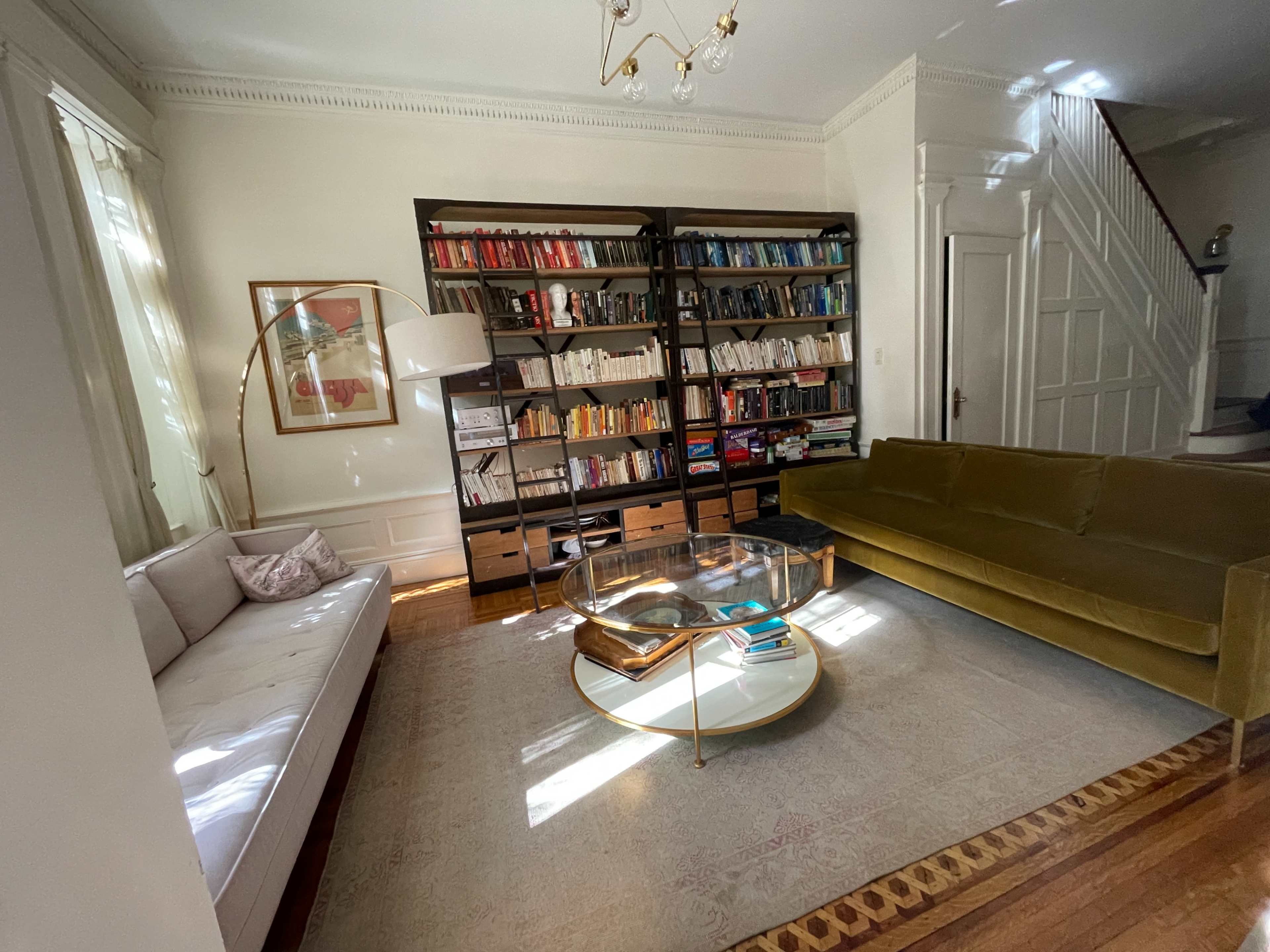 The image shows a cozy living room with a bookshelf filled with books, a glass coffee table, a green velvet sofa, and a white couch.