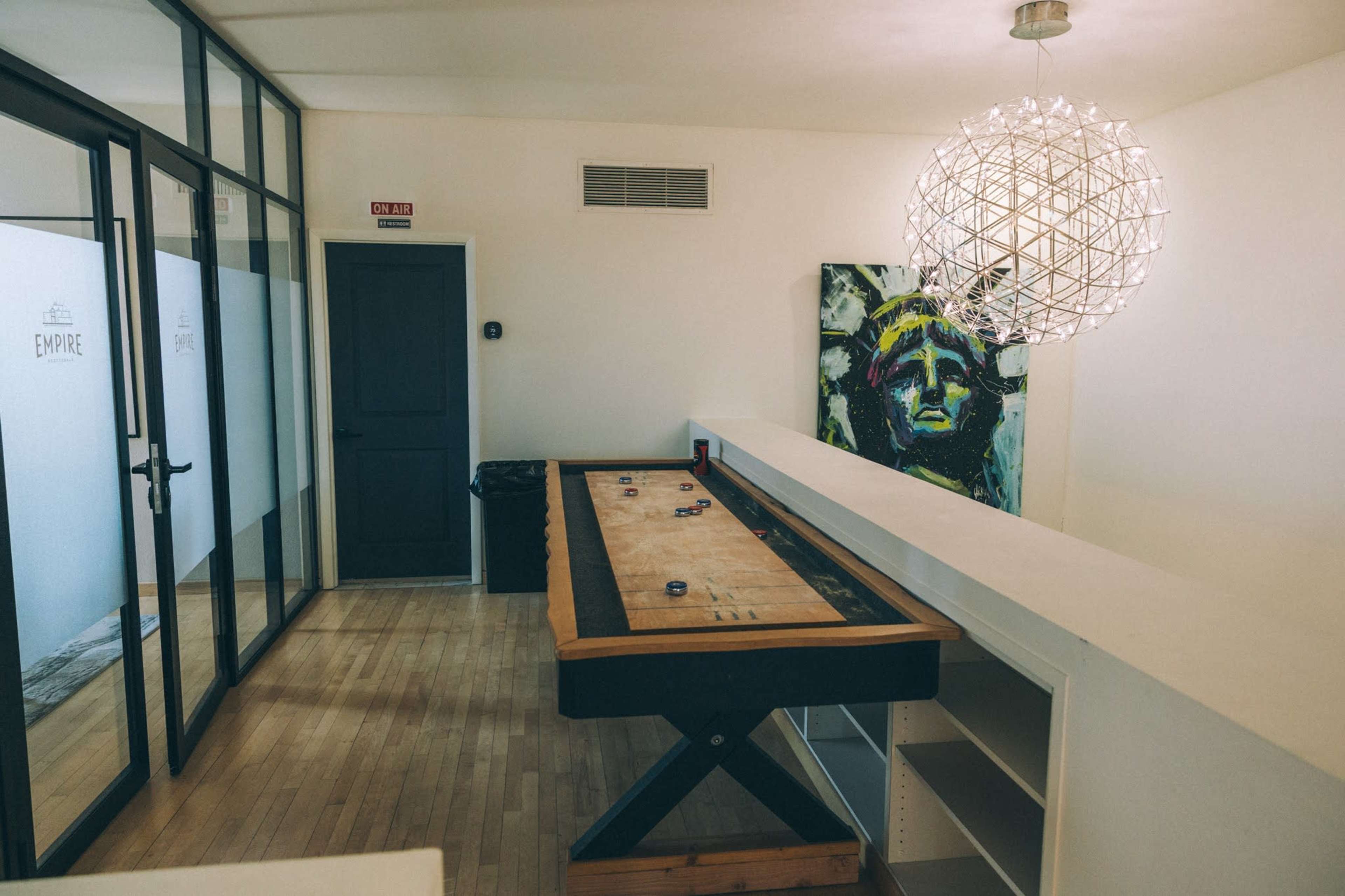 The image shows a modern room with a large shuffleboard table, a minimalist light fixture, and a vibrant painting on the wall.