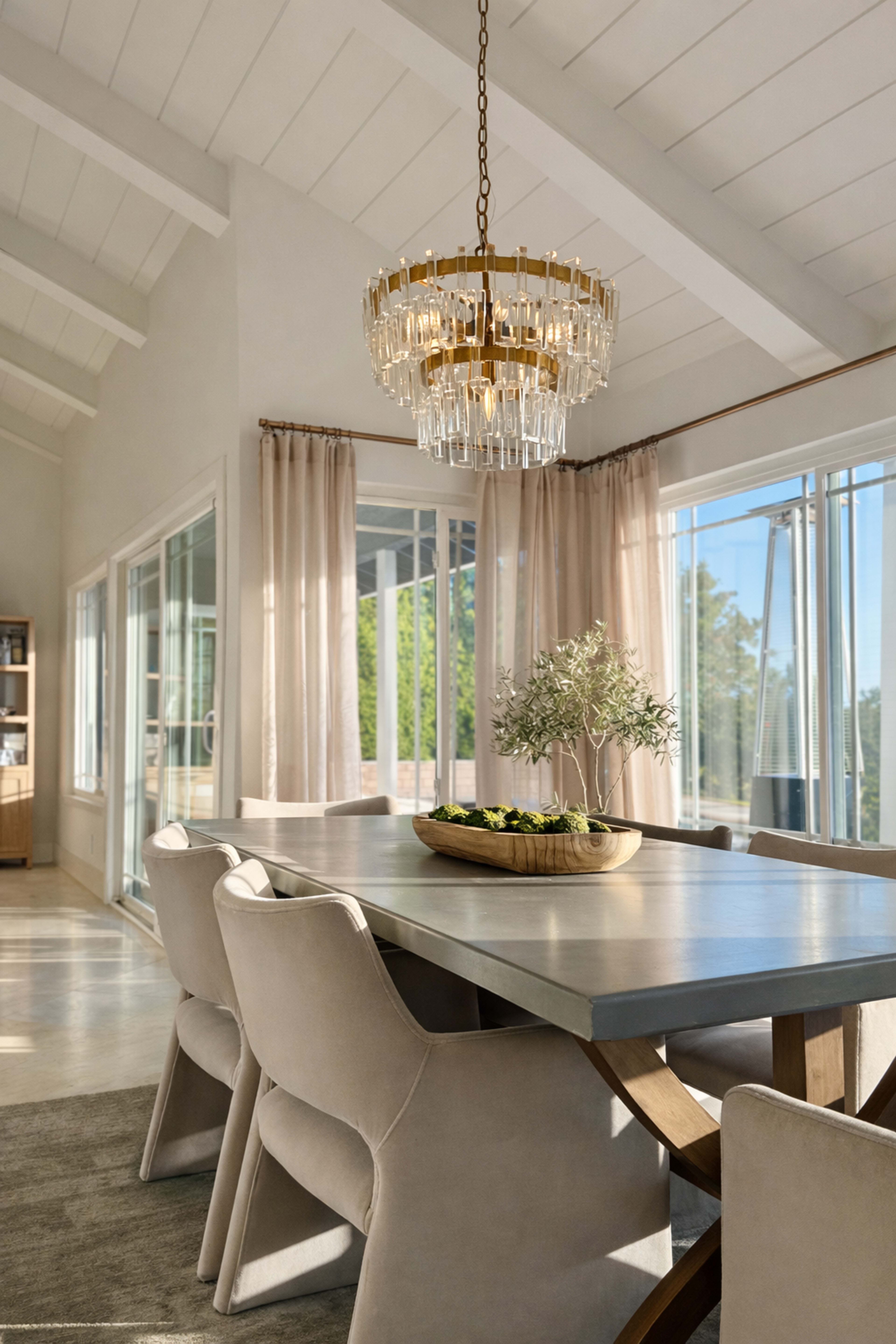 A modern dining room features a large wooden table surrounded by upholstered chairs, with a crystal chandelier hanging overhead and large windows allowing natural light to fill the space.