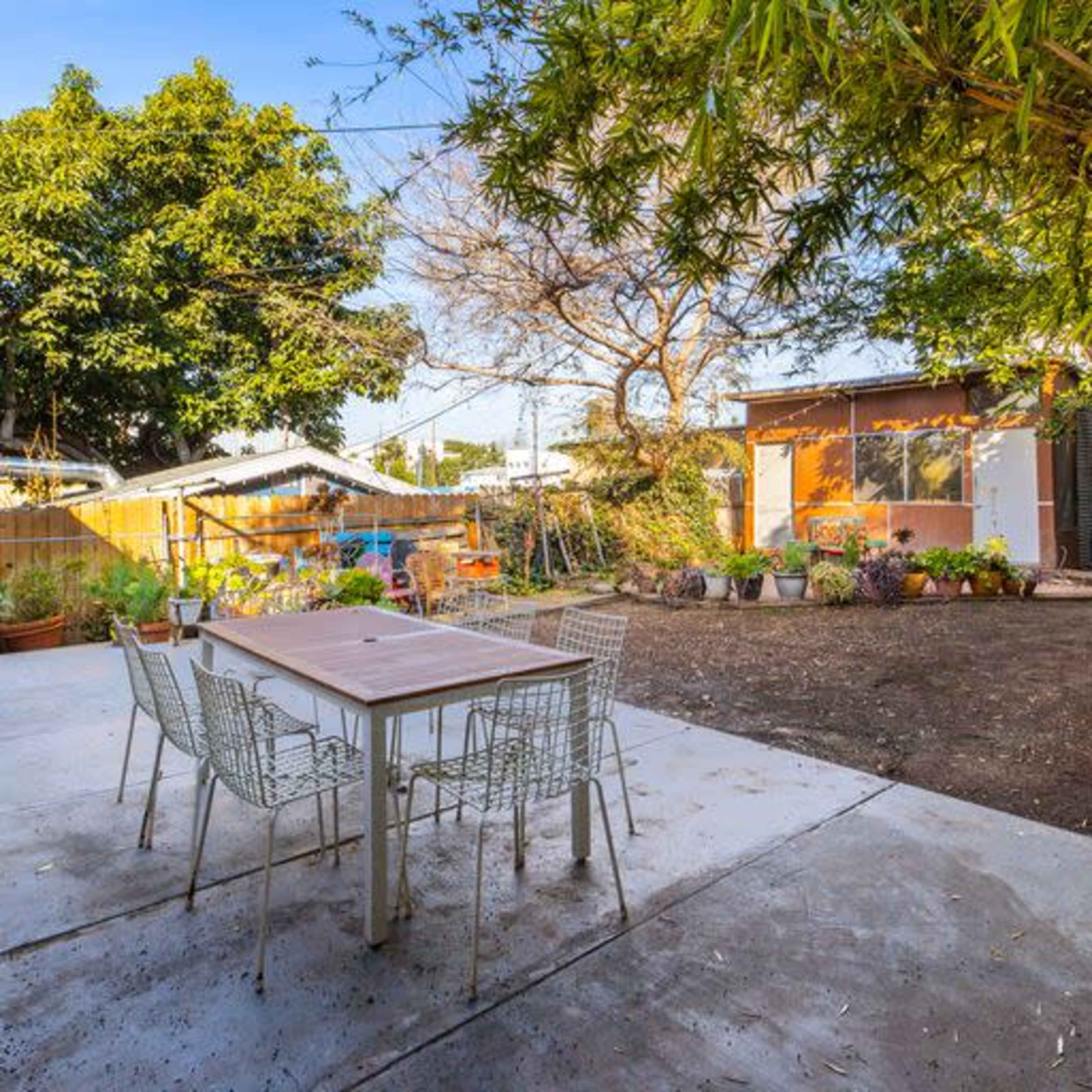 A patio area features a rectangular table surrounded by wire chairs, with trees and a garden in the background.