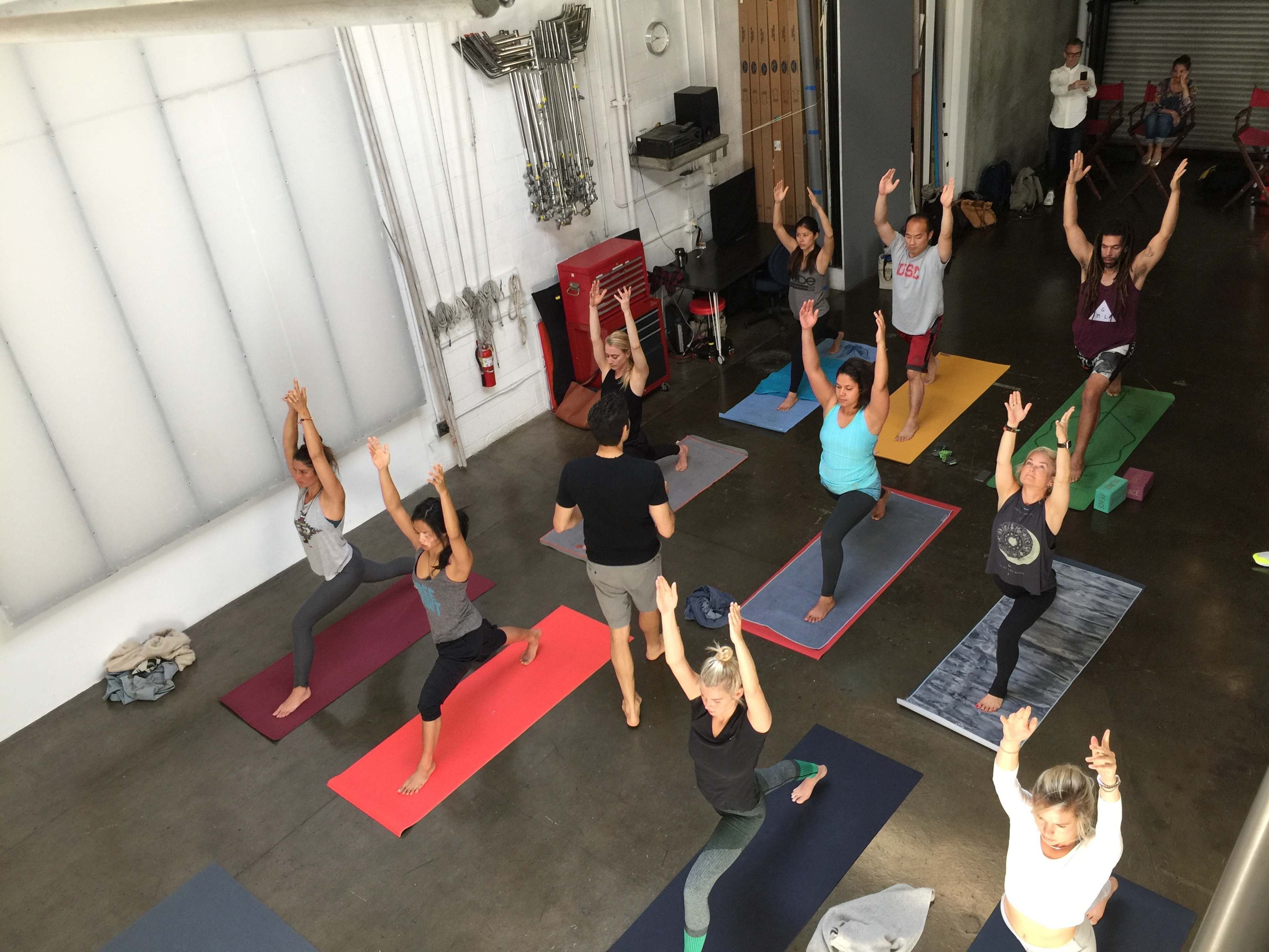 A group of people is practicing yoga in a spacious studio, with mats arranged in rows and various poses being performed.