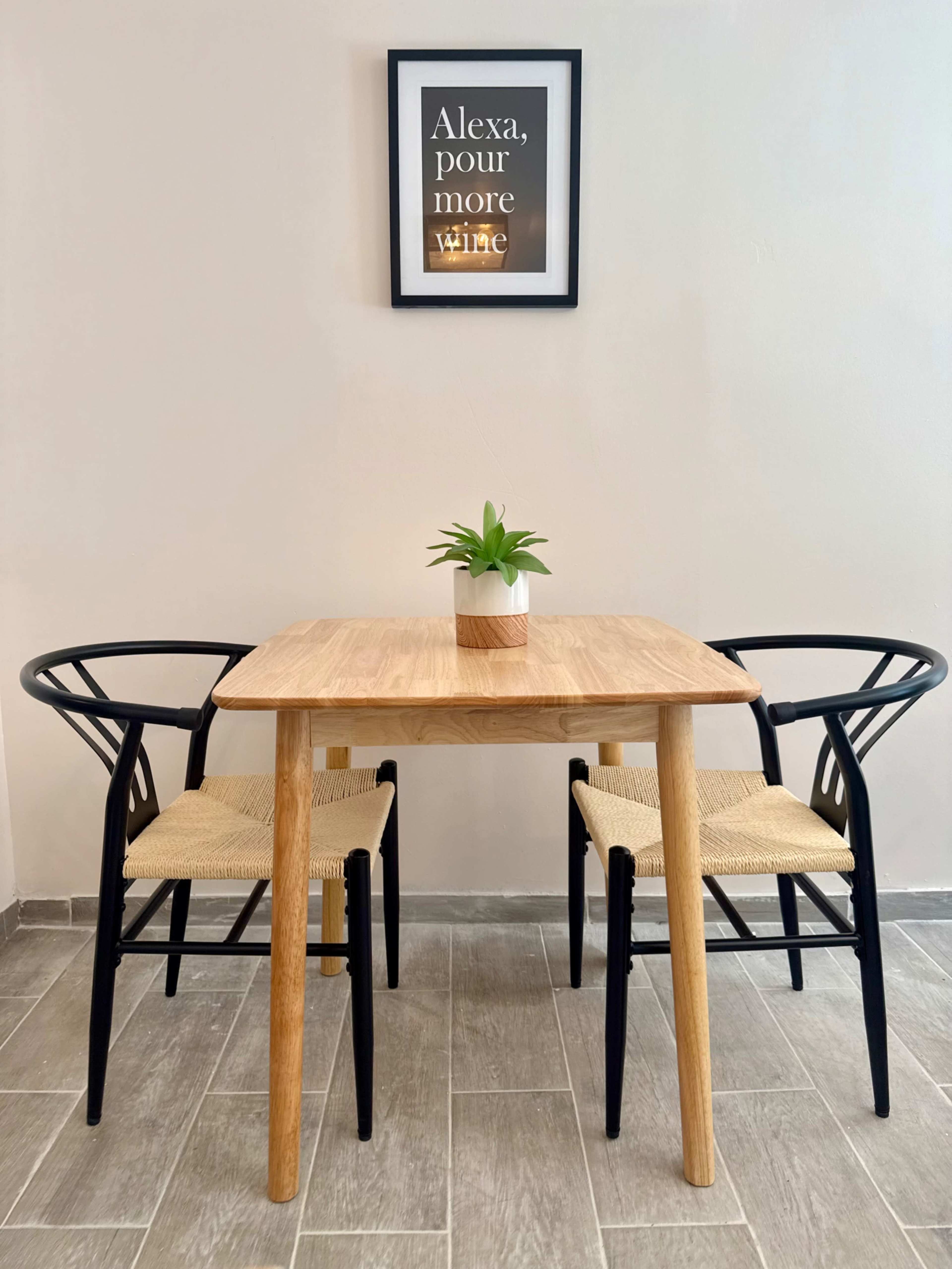 A wooden table with two chairs on either side sits against a wall, featuring a framed print above that reads "Alexa, pour more wine."