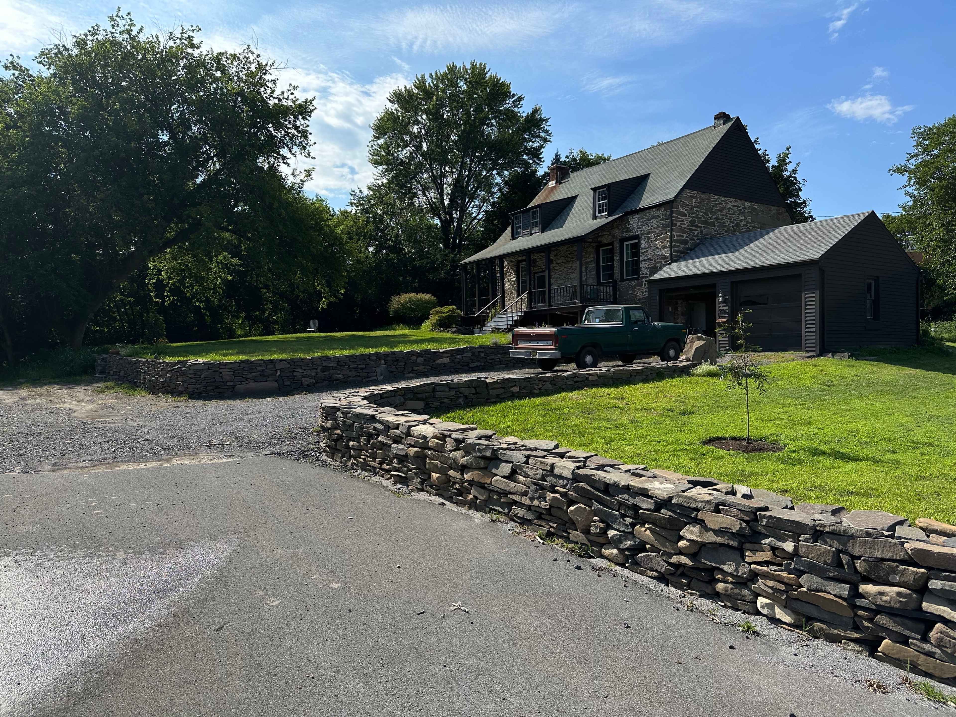 A stone house with a sloped roof is set on a grassy lot with a gravel driveway and a pickup truck parked nearby.