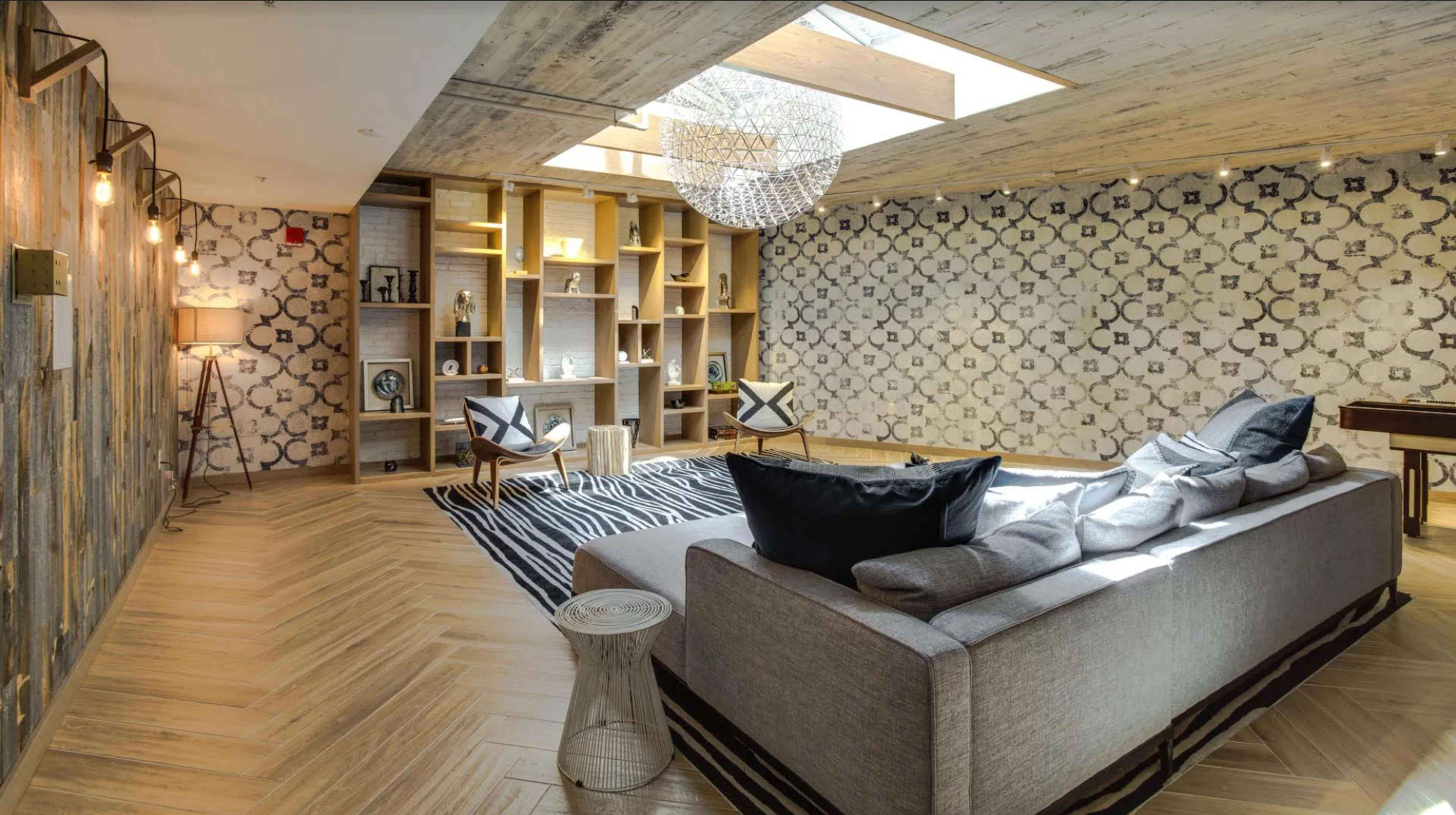 A modern living room featuring a light gray sofa, patterned walls, a geometric chandelier, and wooden flooring with a central area rug.