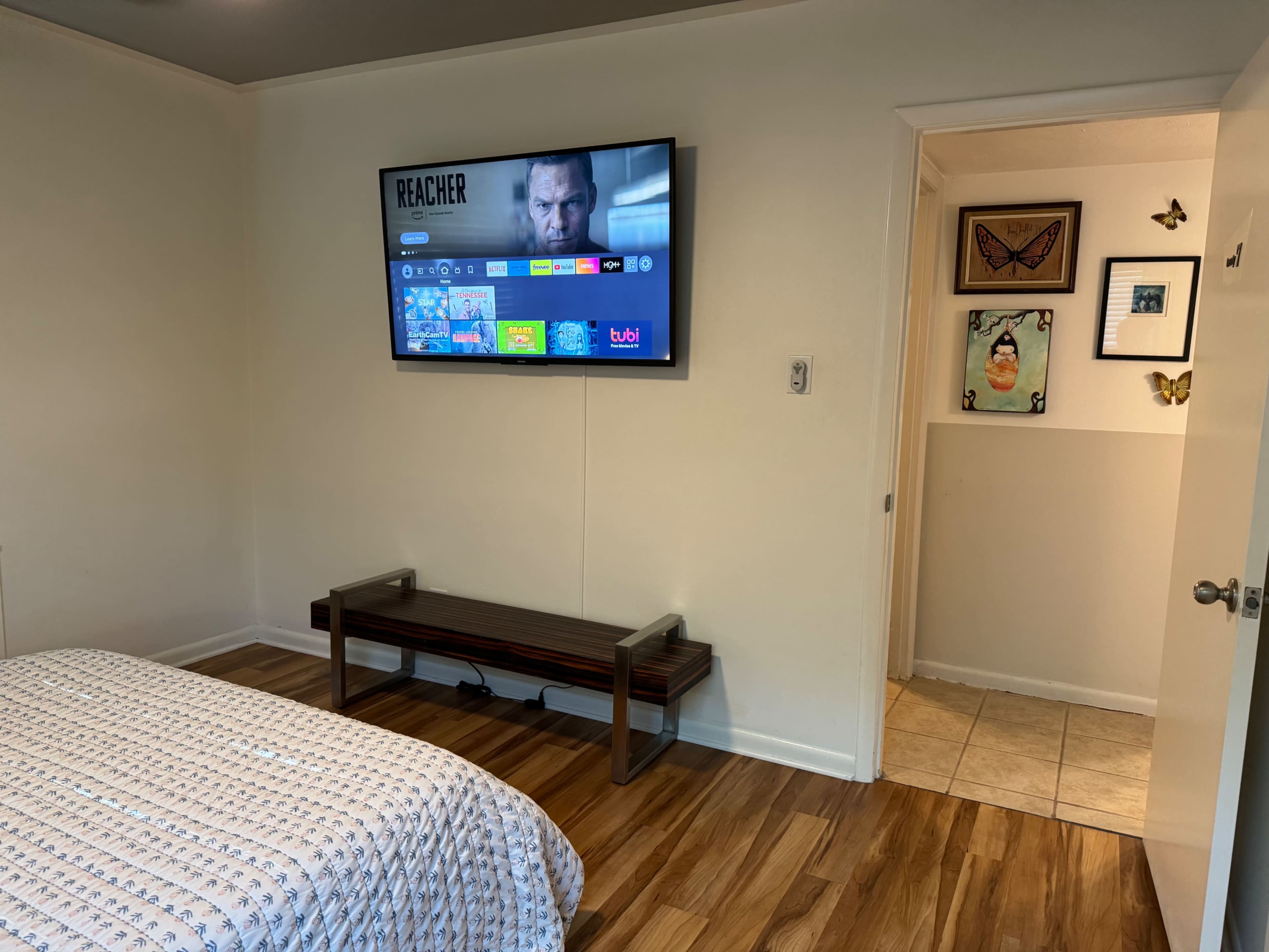 A television is mounted on the wall in a room with a bed and a wooden bench, leading to a doorway that opens into a tiled area.