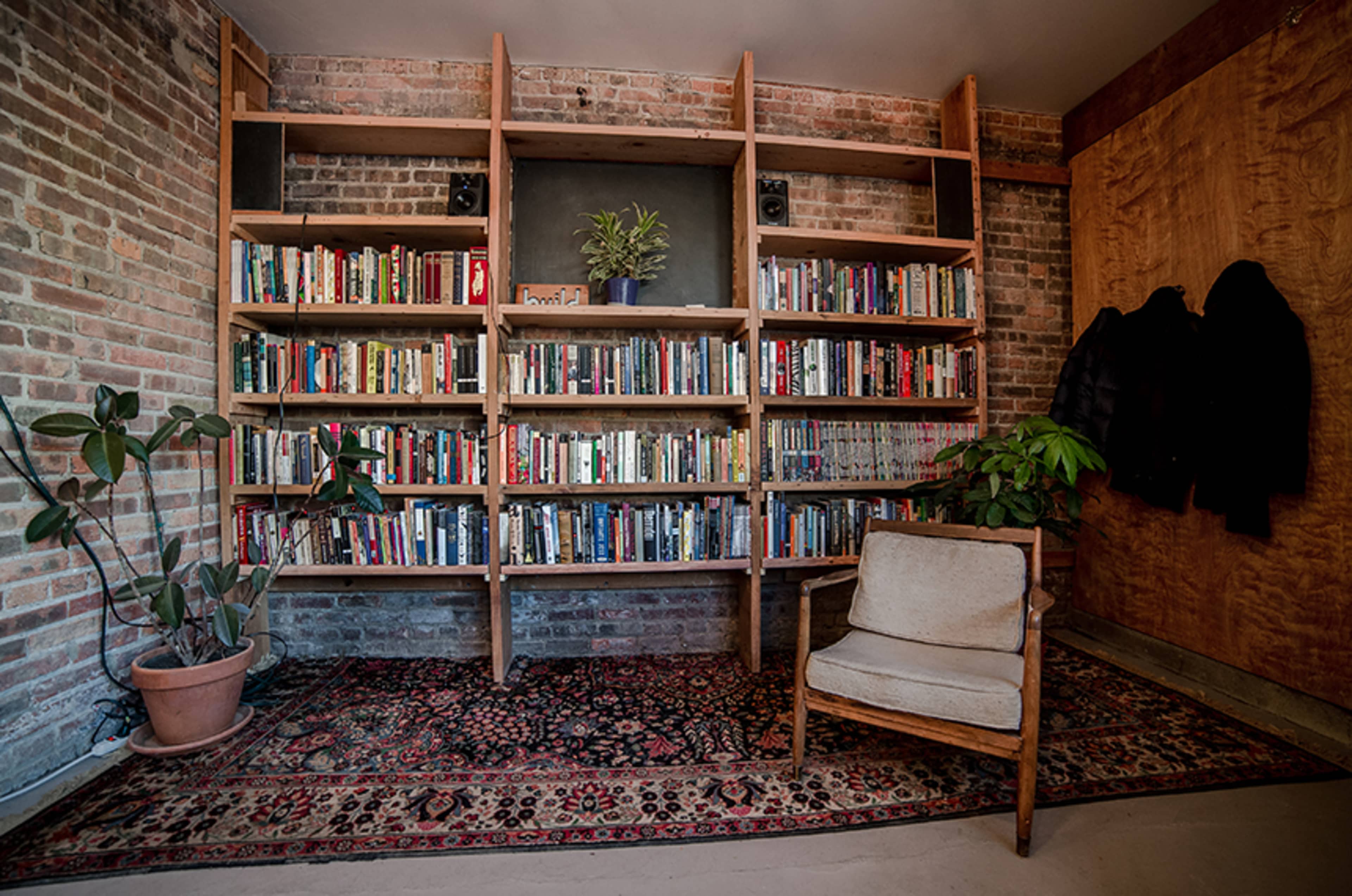 A wooden bookshelf filled with books stands against a brick wall in a cozy room with a patterned rug and a chair.