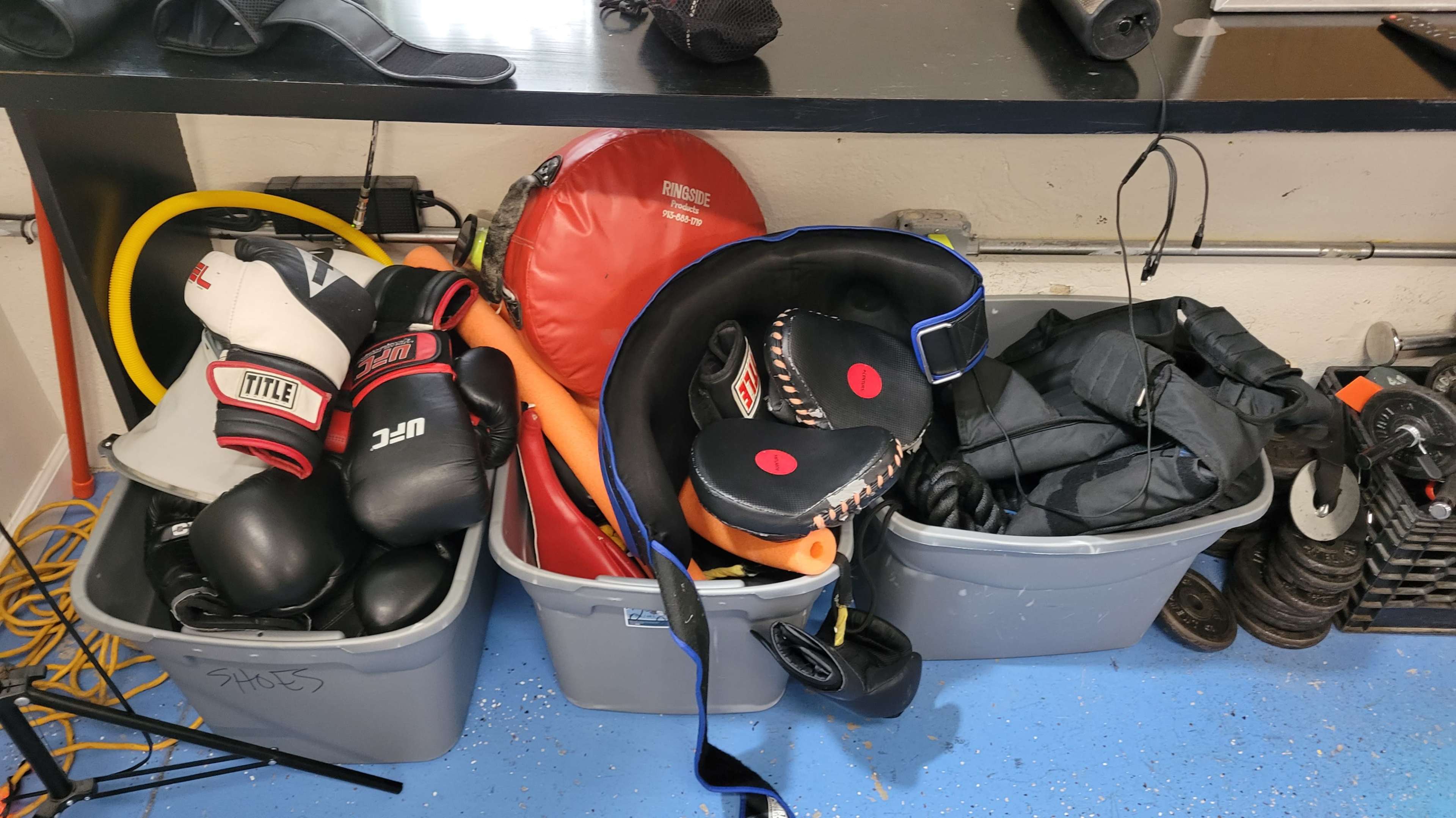 The image shows several storage bins filled with boxing gloves, training pads, resistance bands, and weights piled in a gym setting.