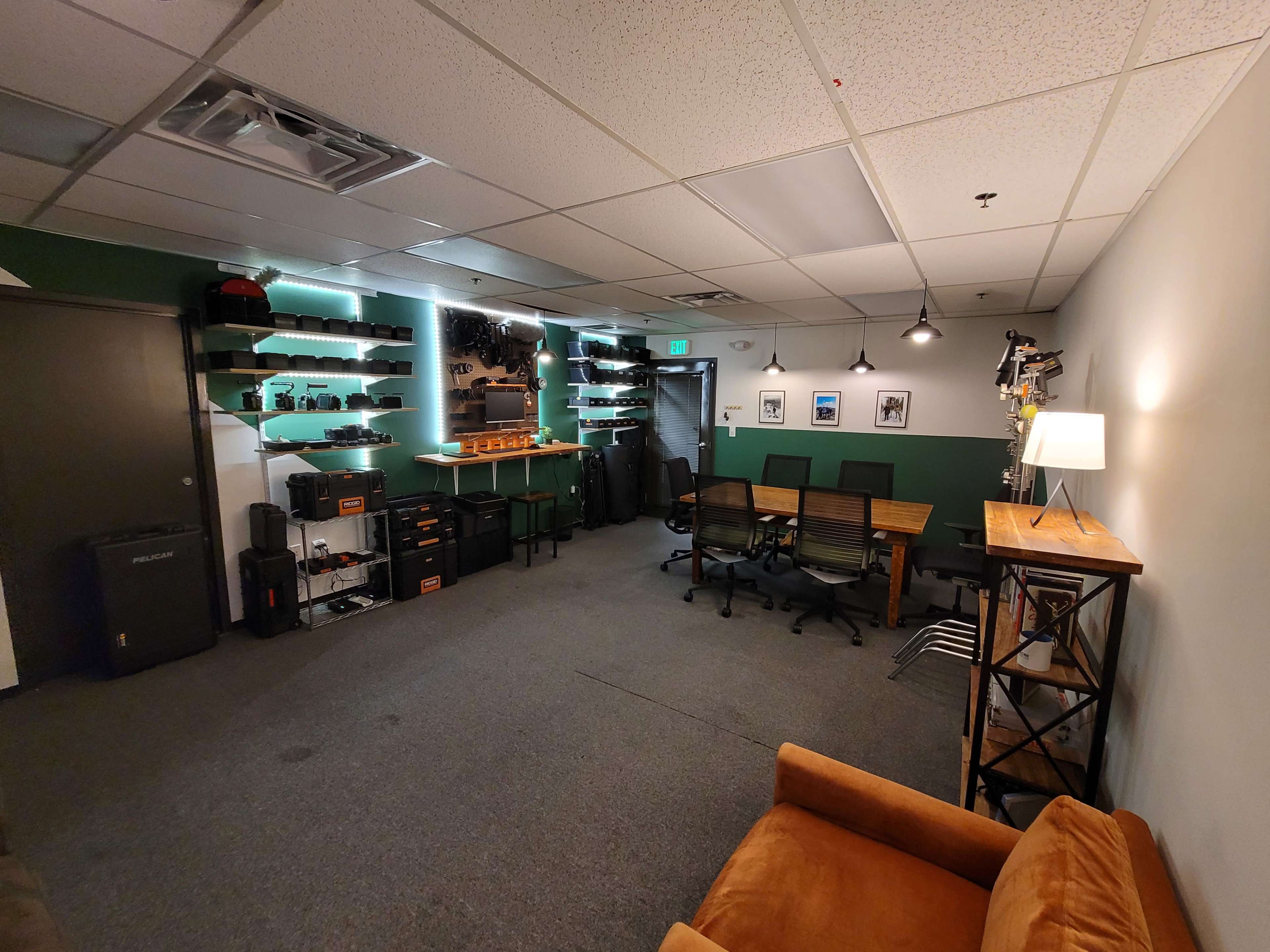 A modern office space with green accent walls, equipped with shelves holding equipment, several chairs around a table, and a cozy seating area featuring an orange couch.