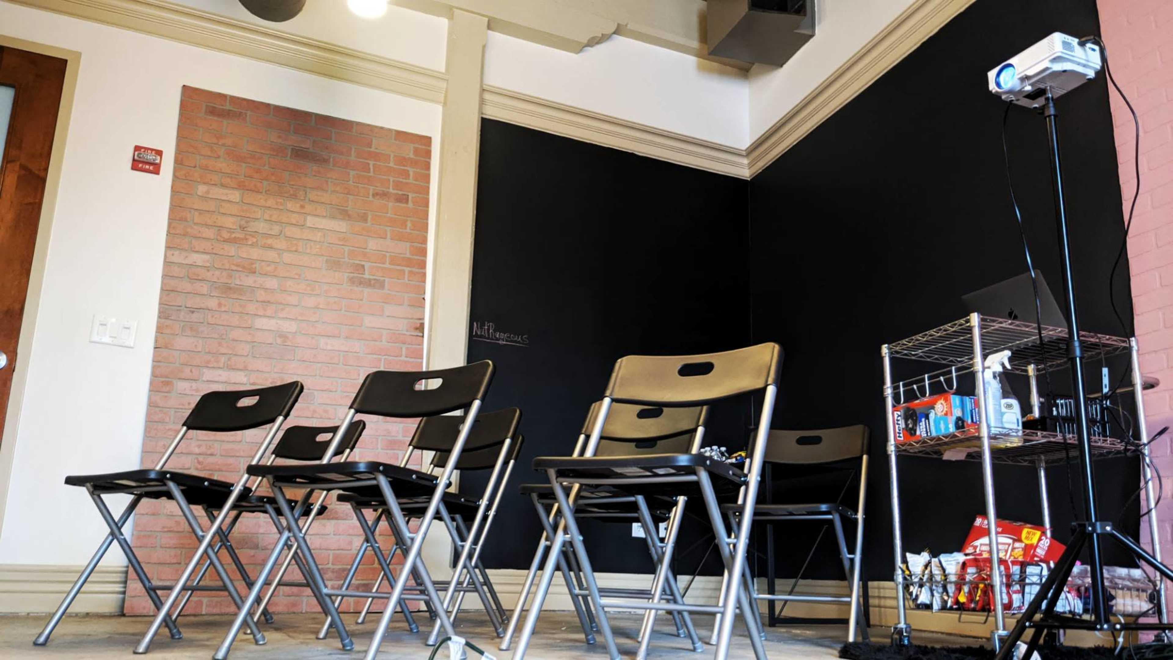 A room with a black chalkboard, several metal folding chairs arranged in a row, and a small cart with various supplies next to a projector.