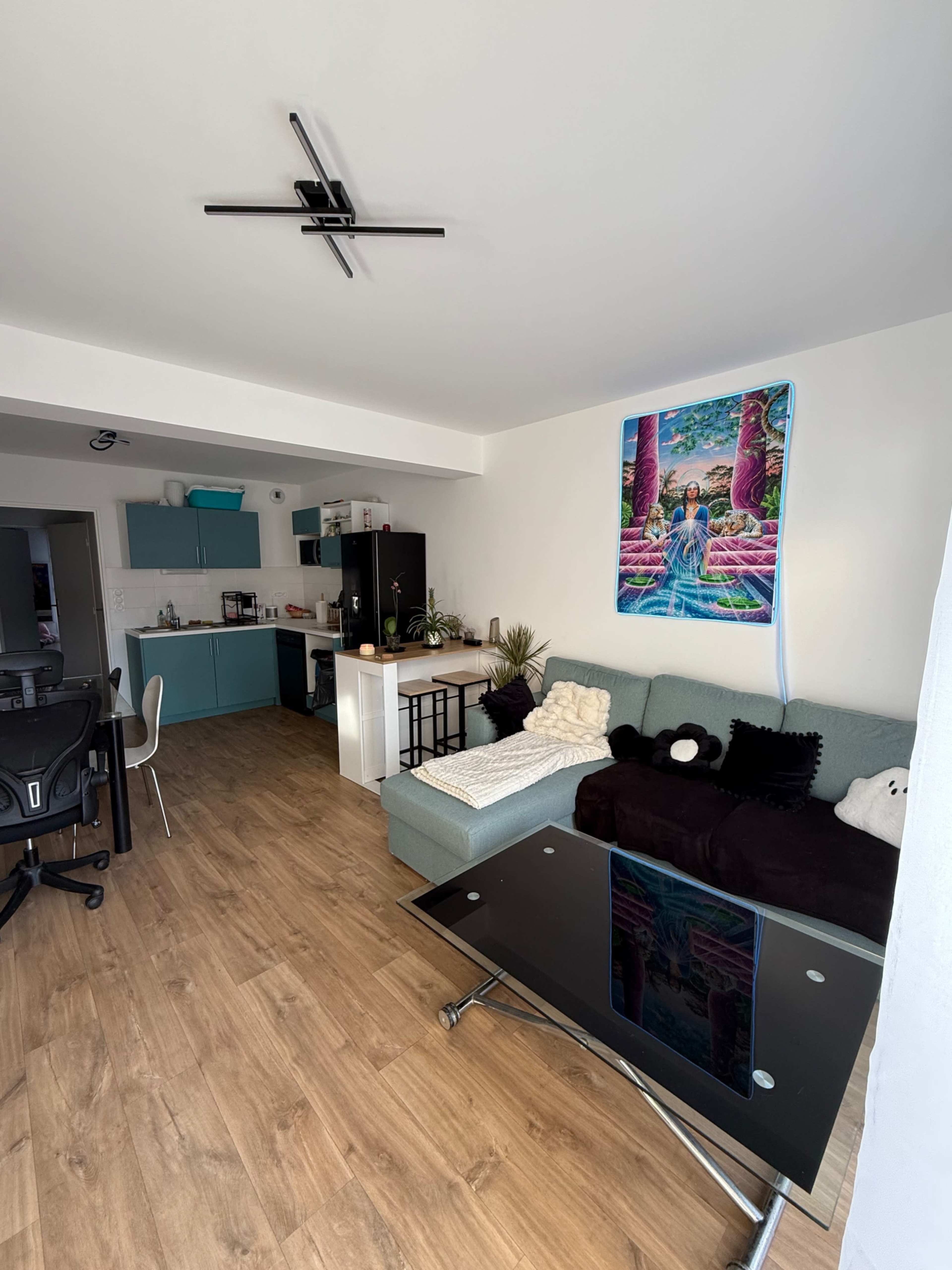 A modern apartment features a living area with a light-colored sofa, a glass coffee table, and a kitchen with teal cabinets in the background.