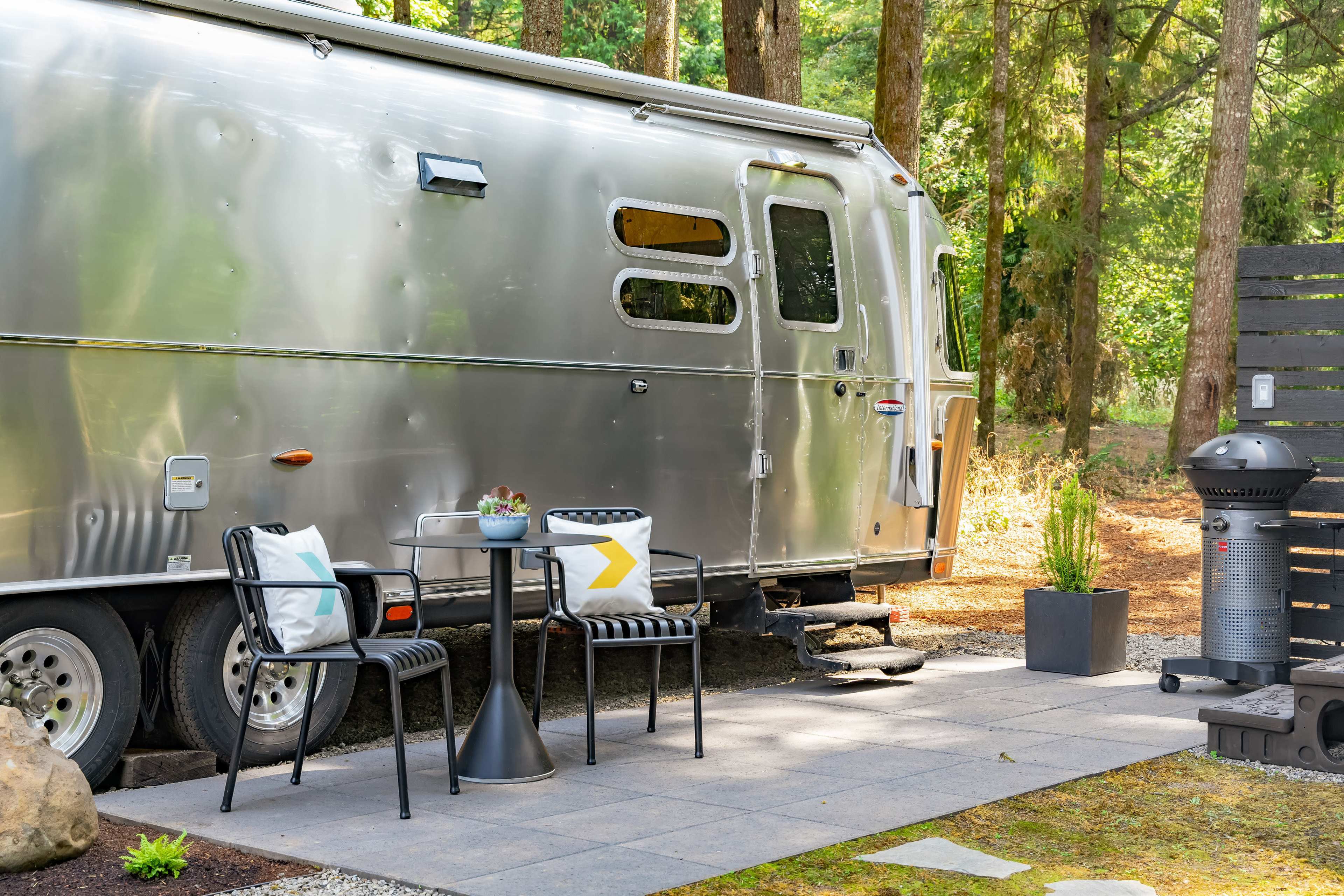 An aluminum Airstream trailer is parked beside a stone patio featuring a small table and chairs, surrounded by trees.