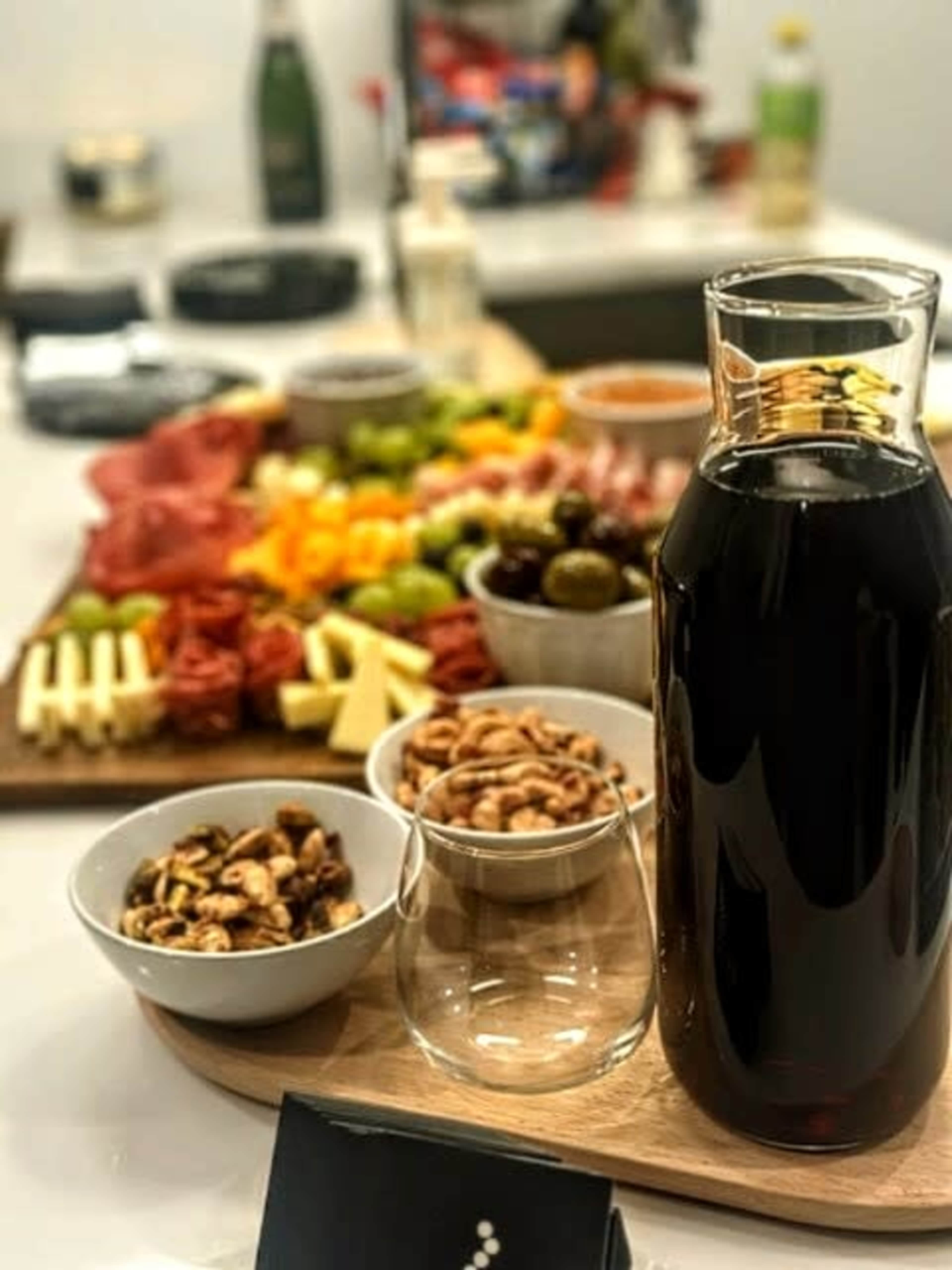 A table is set with a variety of appetizers, including meats, cheeses, fruits, and nuts, alongside a bottle of dark liquid and a small glass.