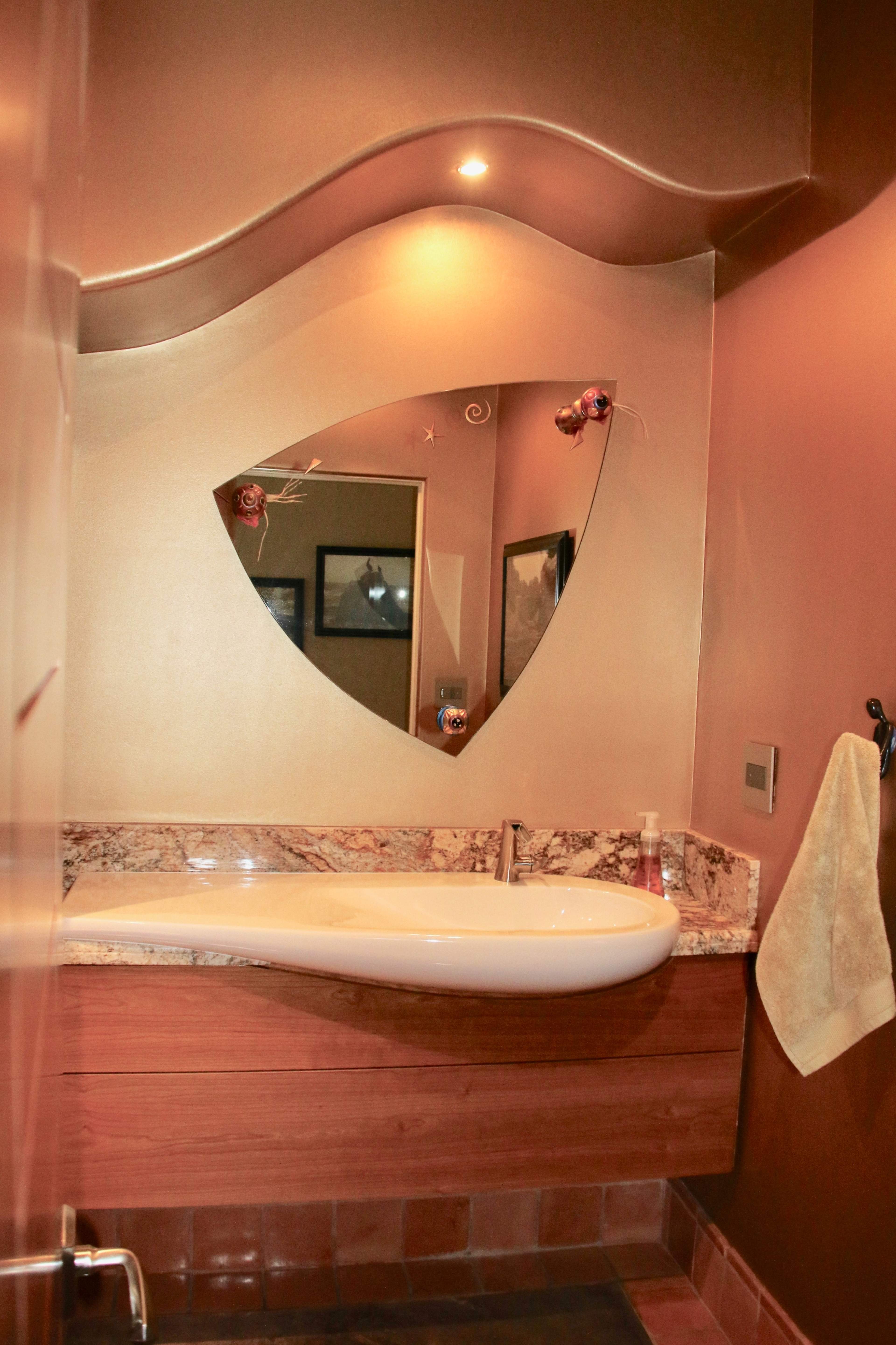 The image shows a modern bathroom featuring a unique wave-shaped mirror above a marble sink, with warm-toned walls and a wooden vanity.