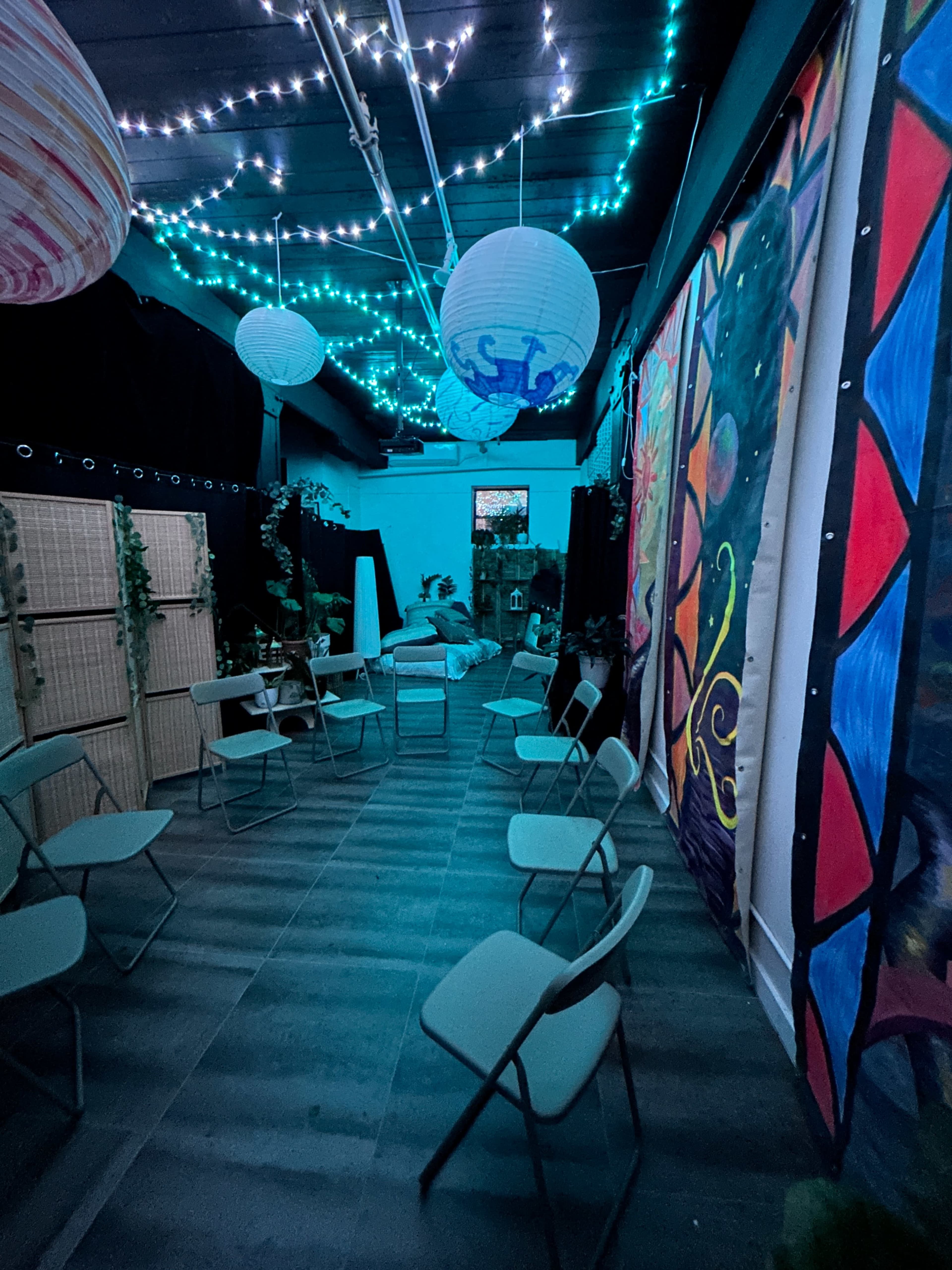 The image shows a dimly lit indoor space with chairs arranged in a circle, colorful lanterns hanging from the ceiling, and vibrant murals on the walls.