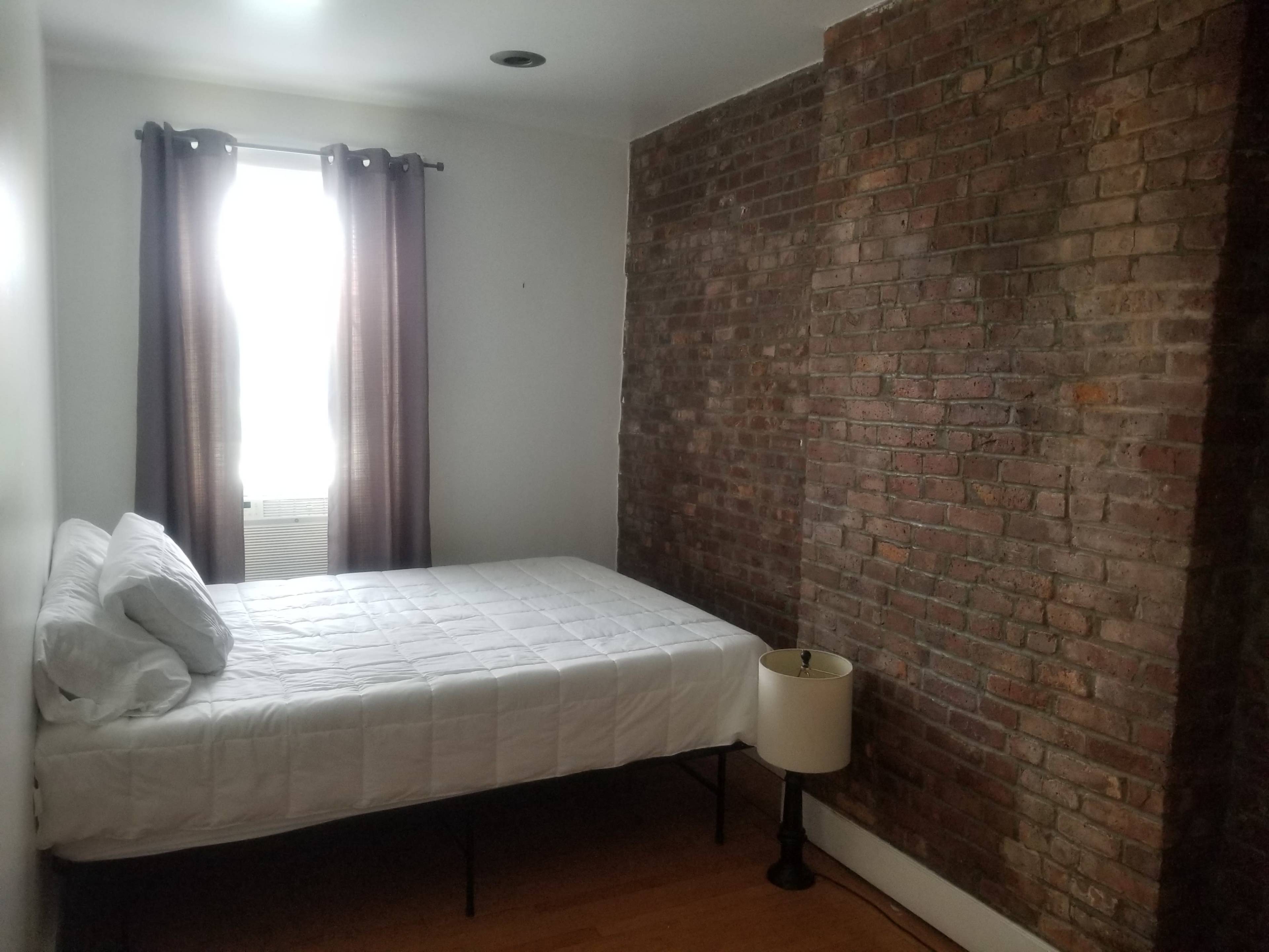 The image shows a simple bedroom with a bed against a rustic brick wall and a lamp beside it.