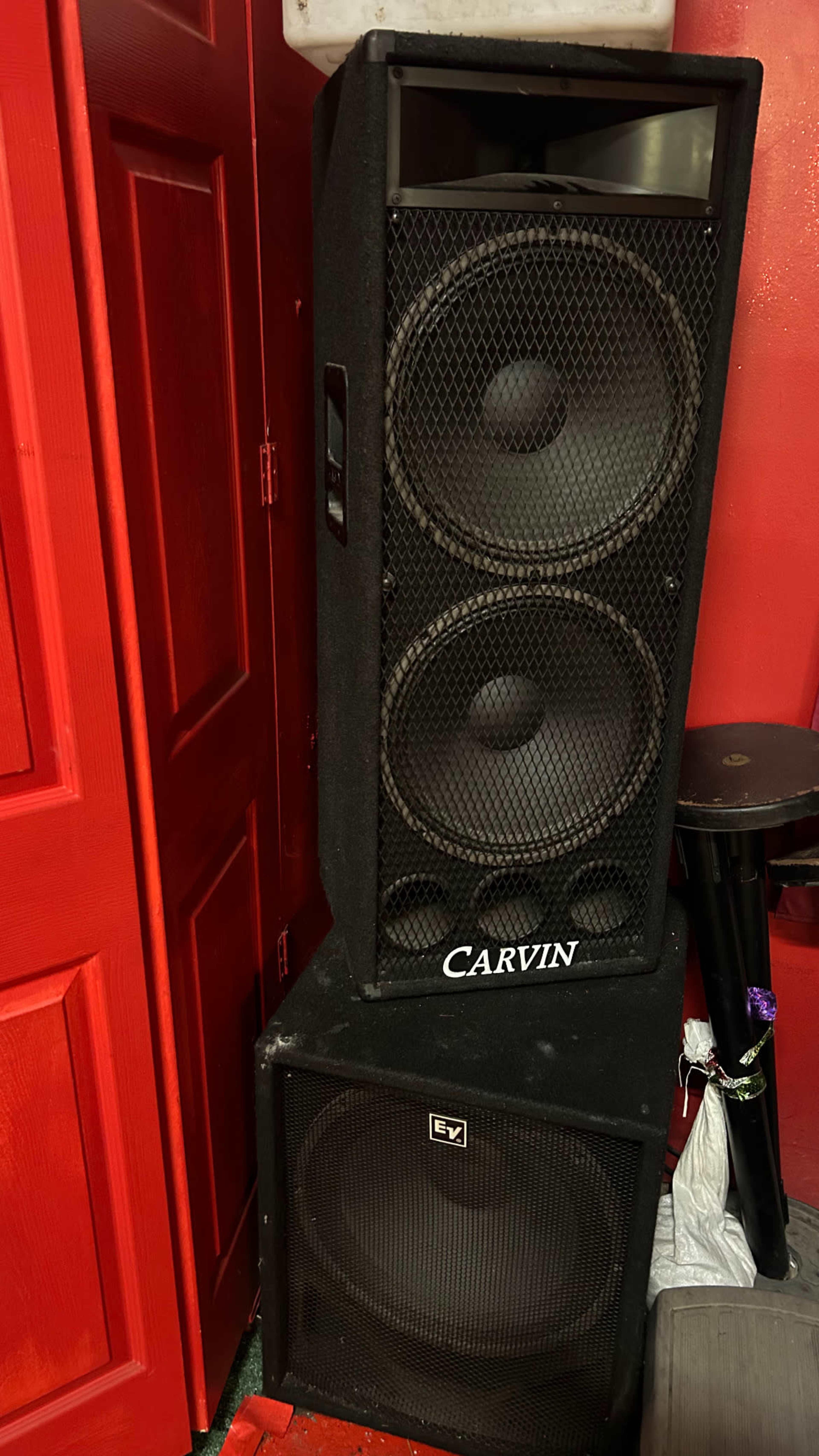 The image shows two large audio speaker units stacked against a red wall.