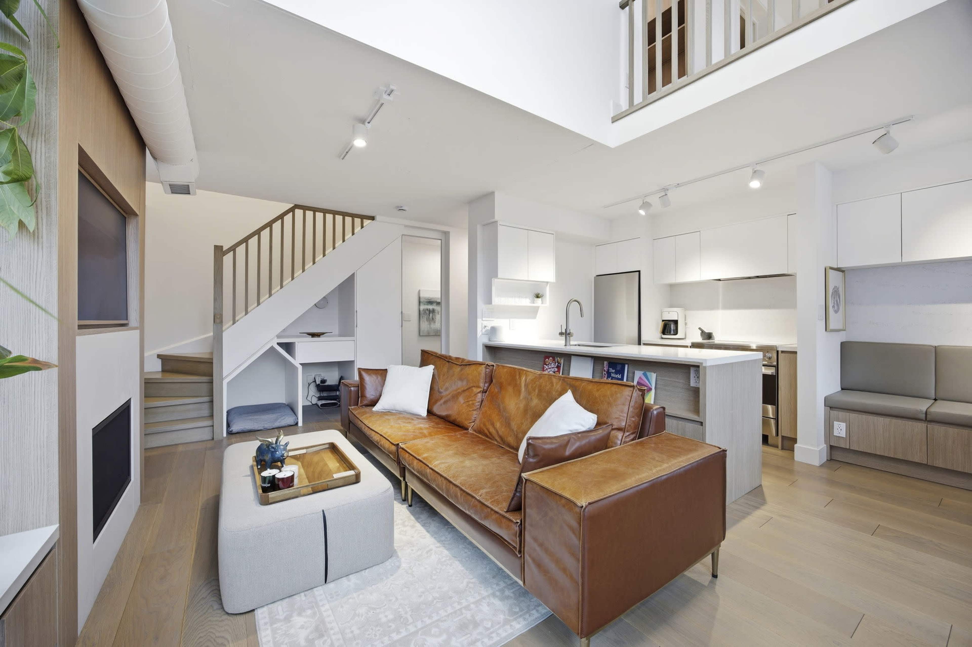 The image shows a modern living space with a leather sofa, a coffee table, and a staircase leading to an upper level, surrounded by light-colored walls and functional furniture.