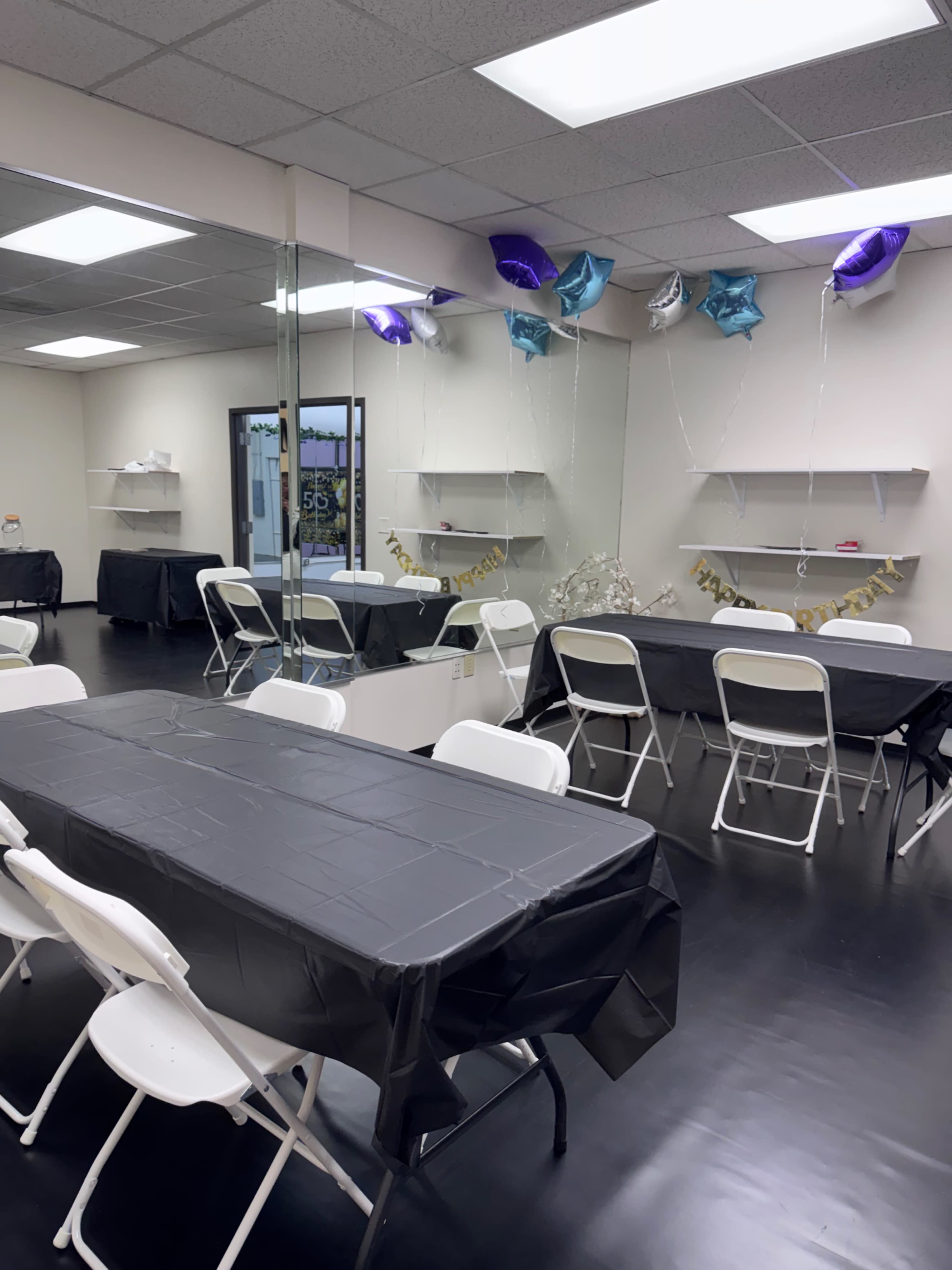 The image shows a spacious room set up for an event, featuring several tables covered with black tablecloths and decorated with purple and silver balloons.