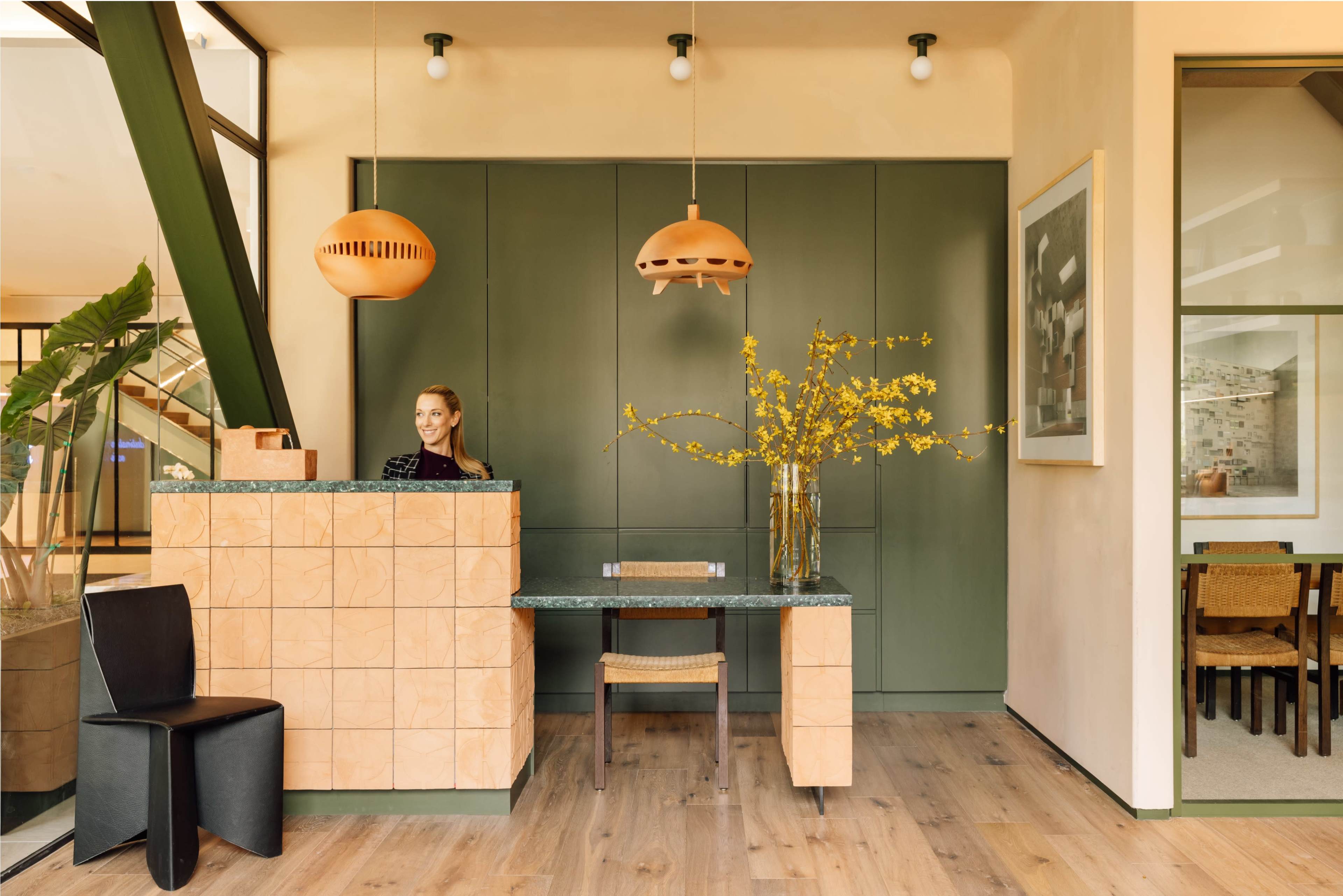 The image shows a modern reception area with a wood and green reception desk, a vase of yellow flowers, and contemporary furnishings.