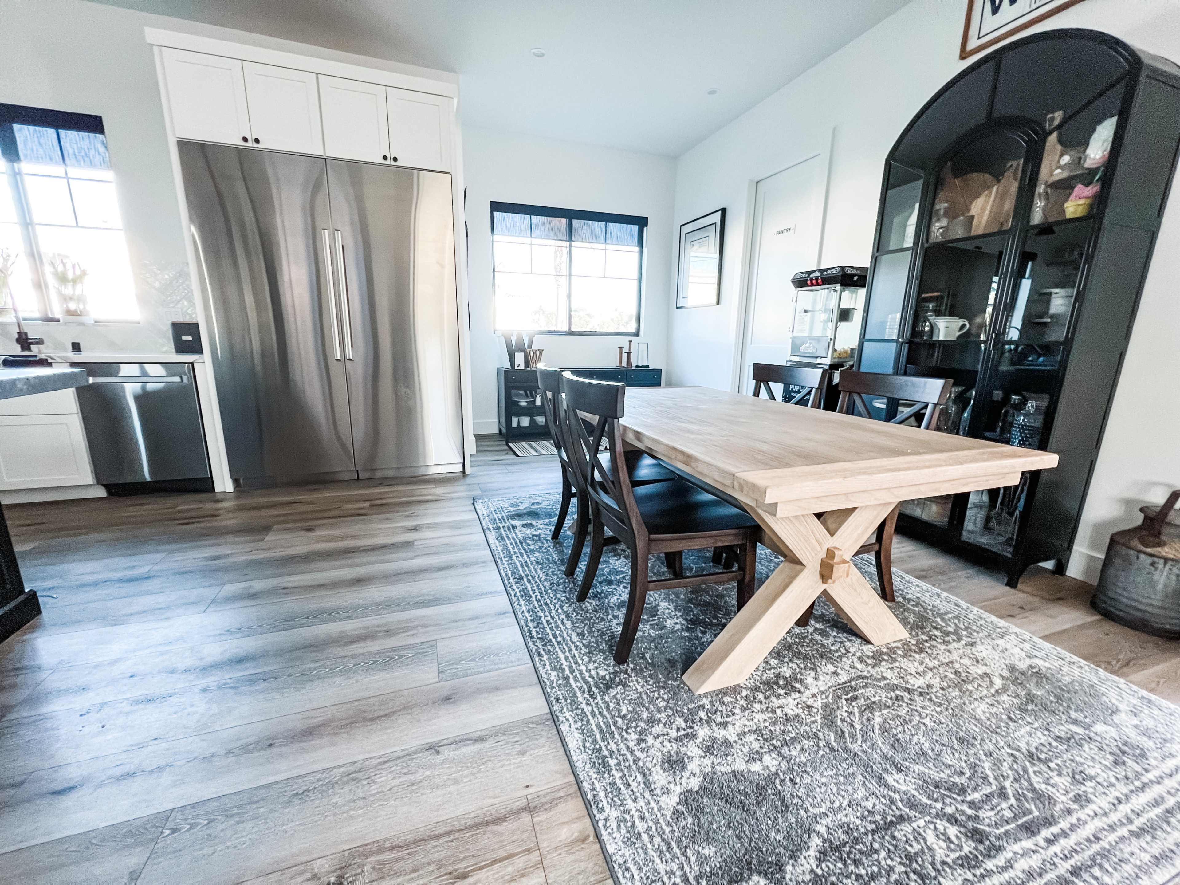 A modern kitchen and dining area featuring a wooden dining table with cross-leg supports, black chairs, and a stainless steel refrigerator.