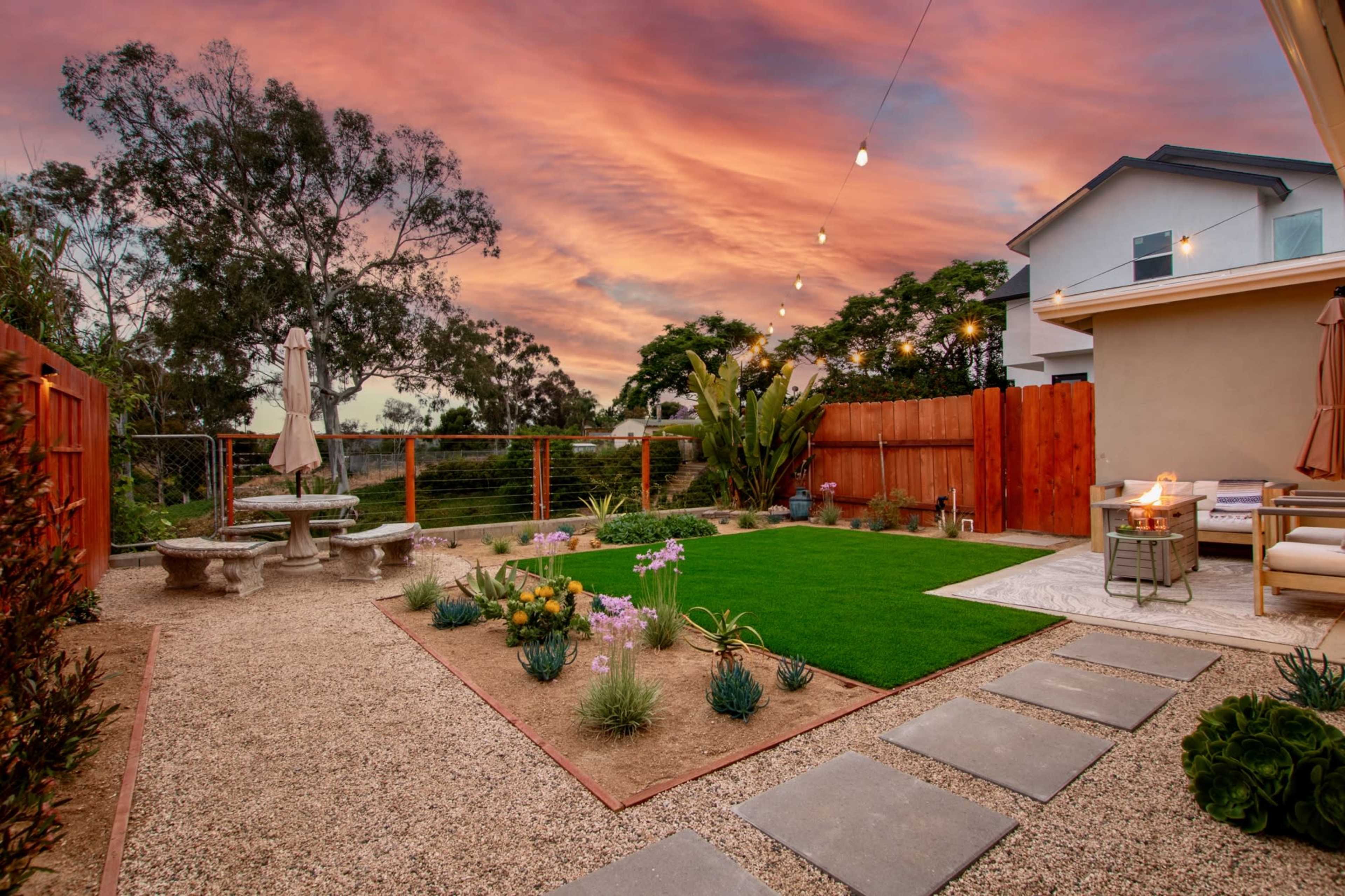 A landscaped backyard with a stone patio, grass area, colorful flowers, and a sunset sky illuminated by string lights.