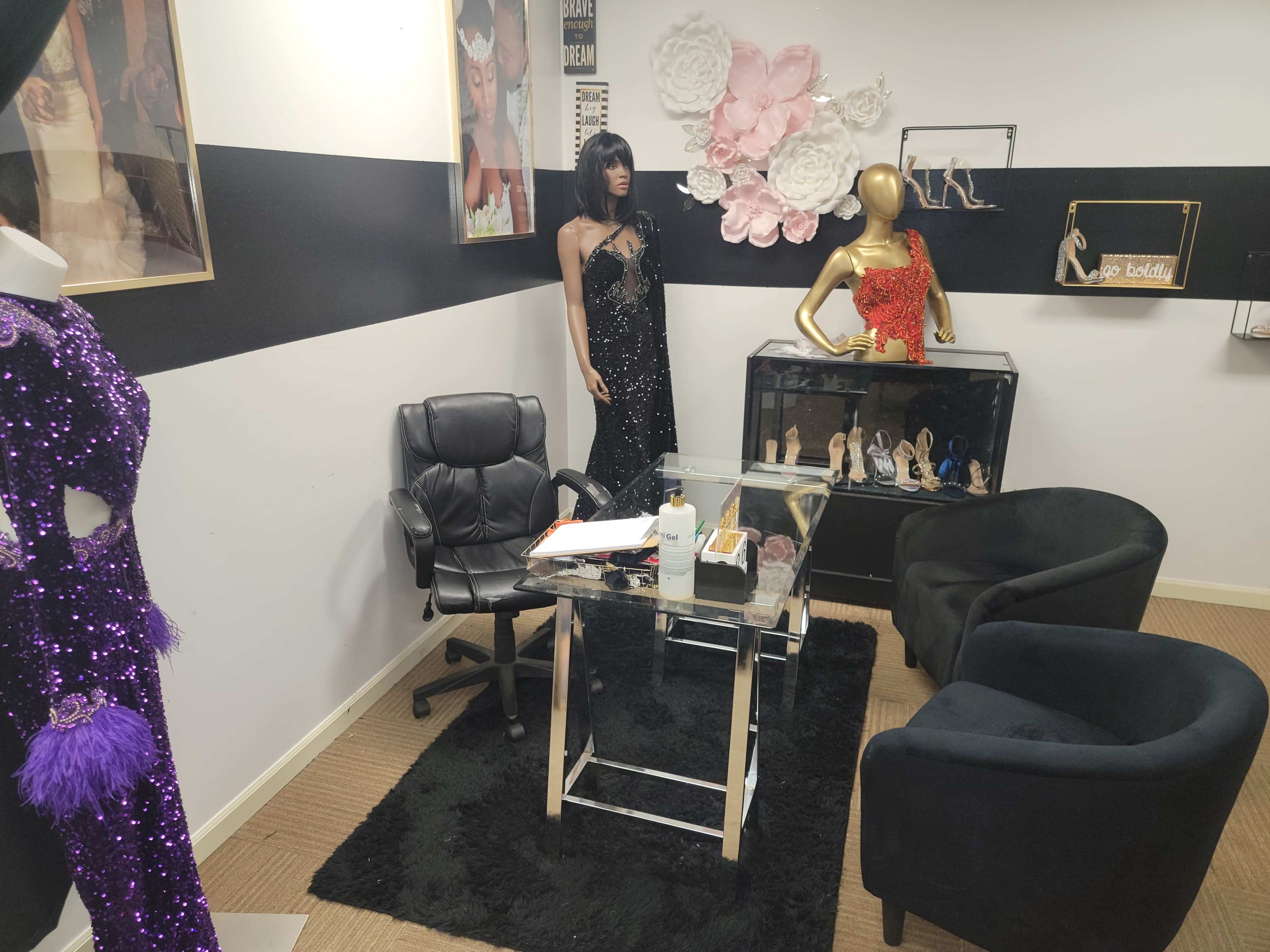 A boutique display featuring evening gowns, a glass table with chairs, and decorative elements such as floral arrangements and high-heeled shoes.