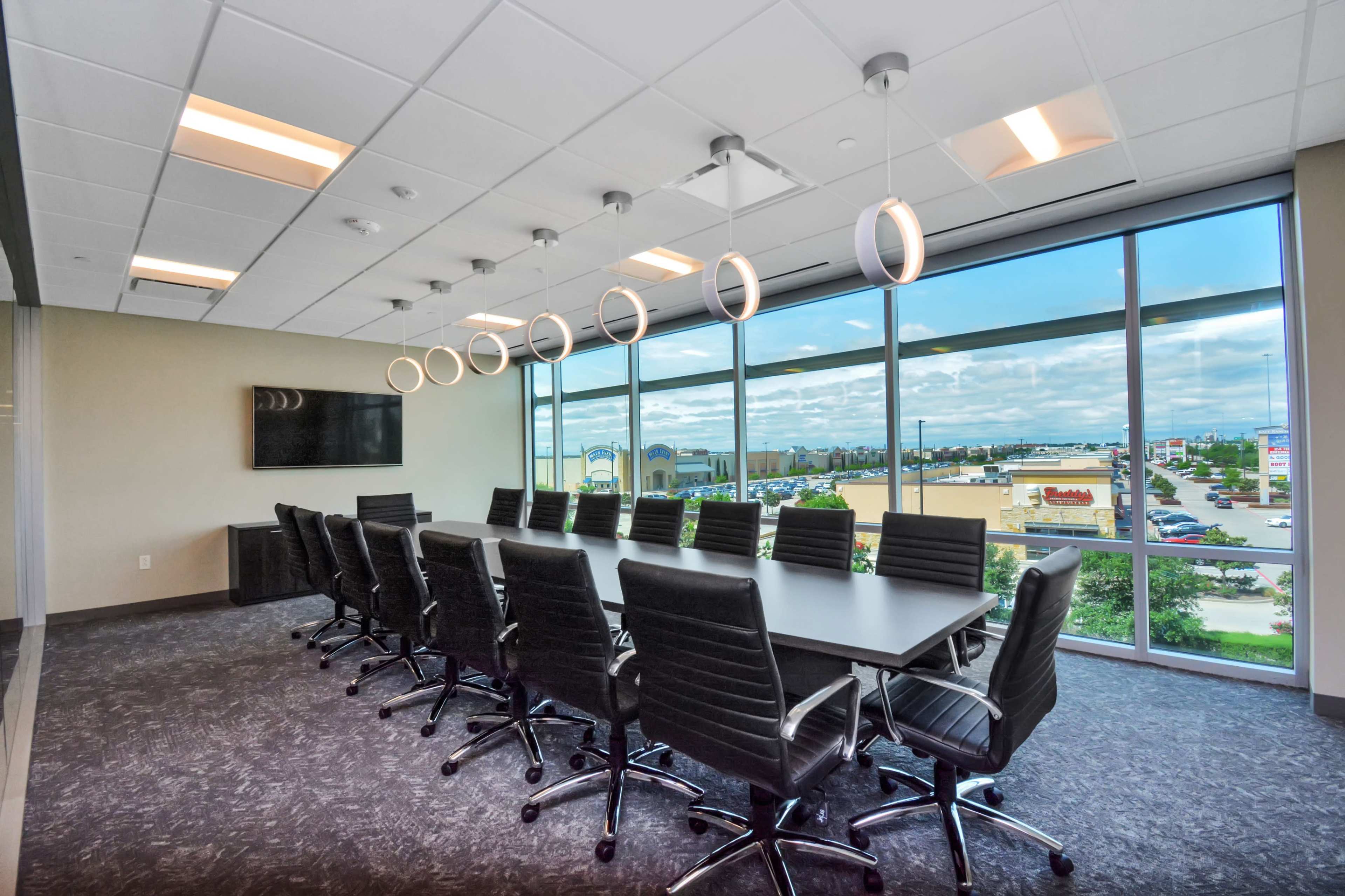 A large, modern conference room features a long black table surrounded by ergonomic chairs, with floor-to-ceiling windows providing a view of the outside.