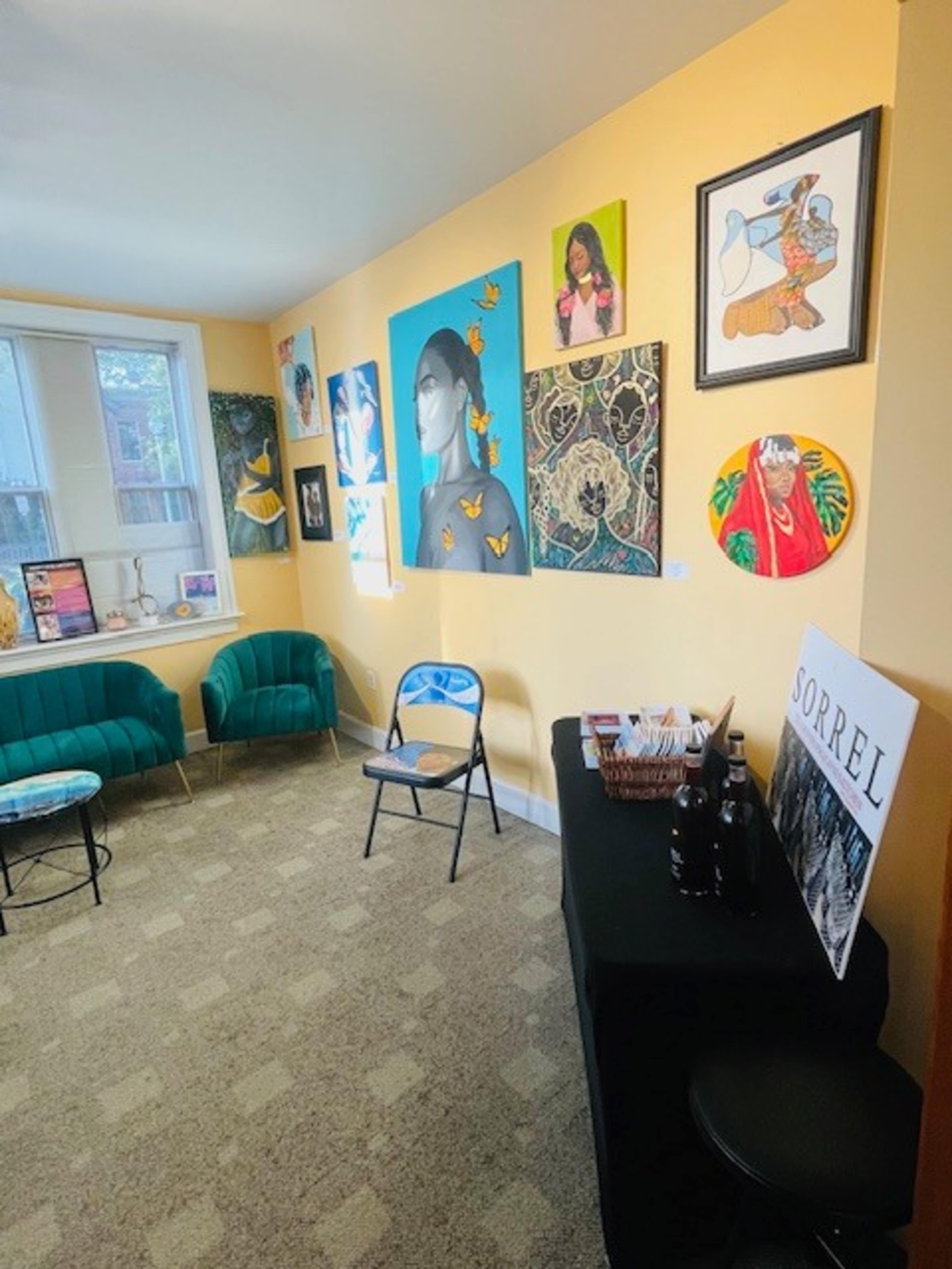 The room features a yellow wall adorned with various colorful paintings, a small round table, teal chairs, and a black table displaying bottles and a sign labeled "SORREL."