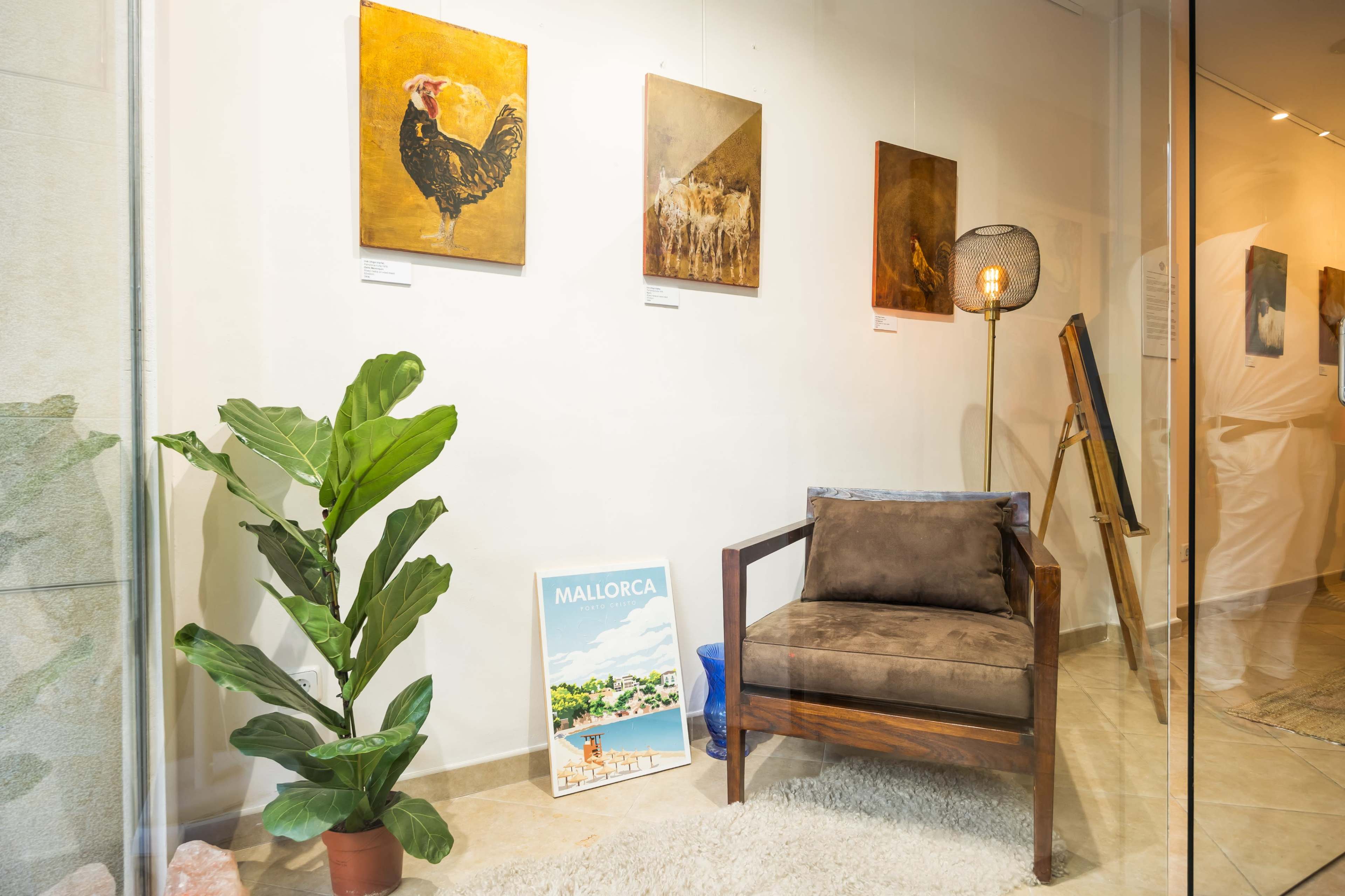 The image shows an art gallery interior featuring paintings on the wall, a brown armchair, a potted plant, and a poster of Mallorca on the floor.