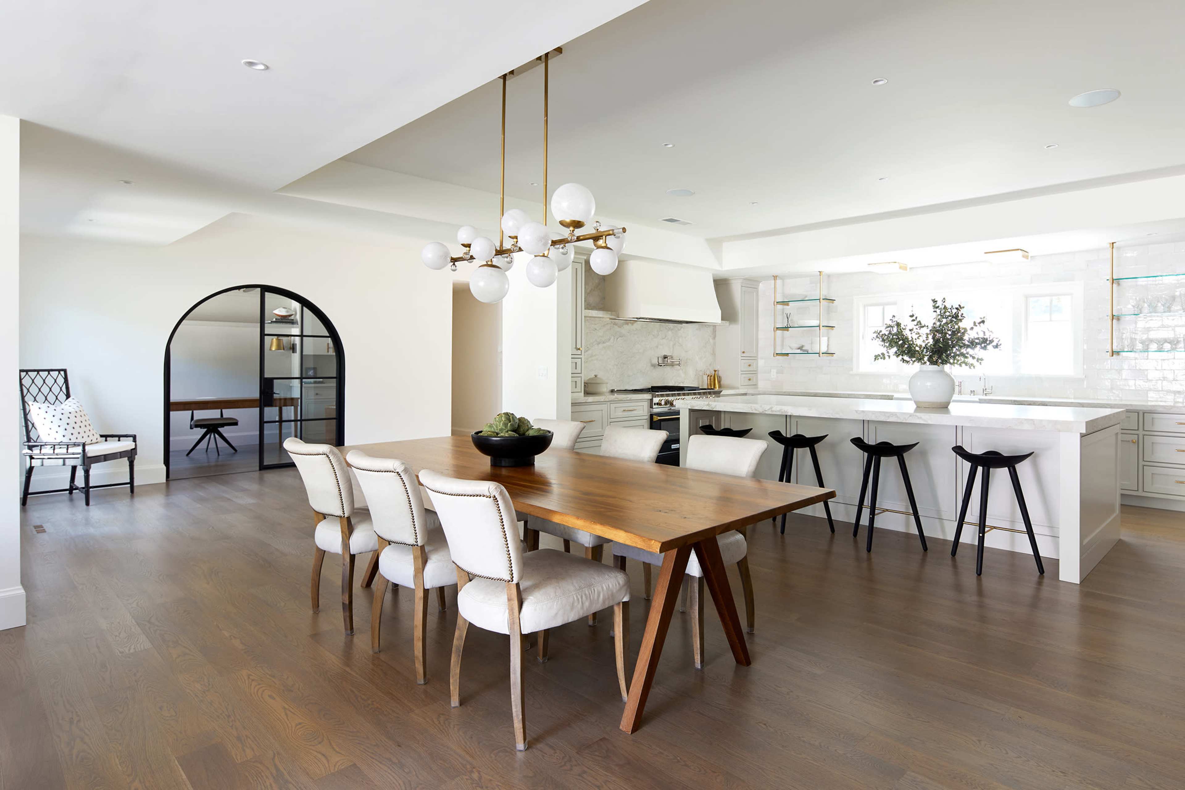 A modern dining area with a wooden table surrounded by six upholstered chairs and a large kitchen in the background, complete with white cabinetry and a sleek island.