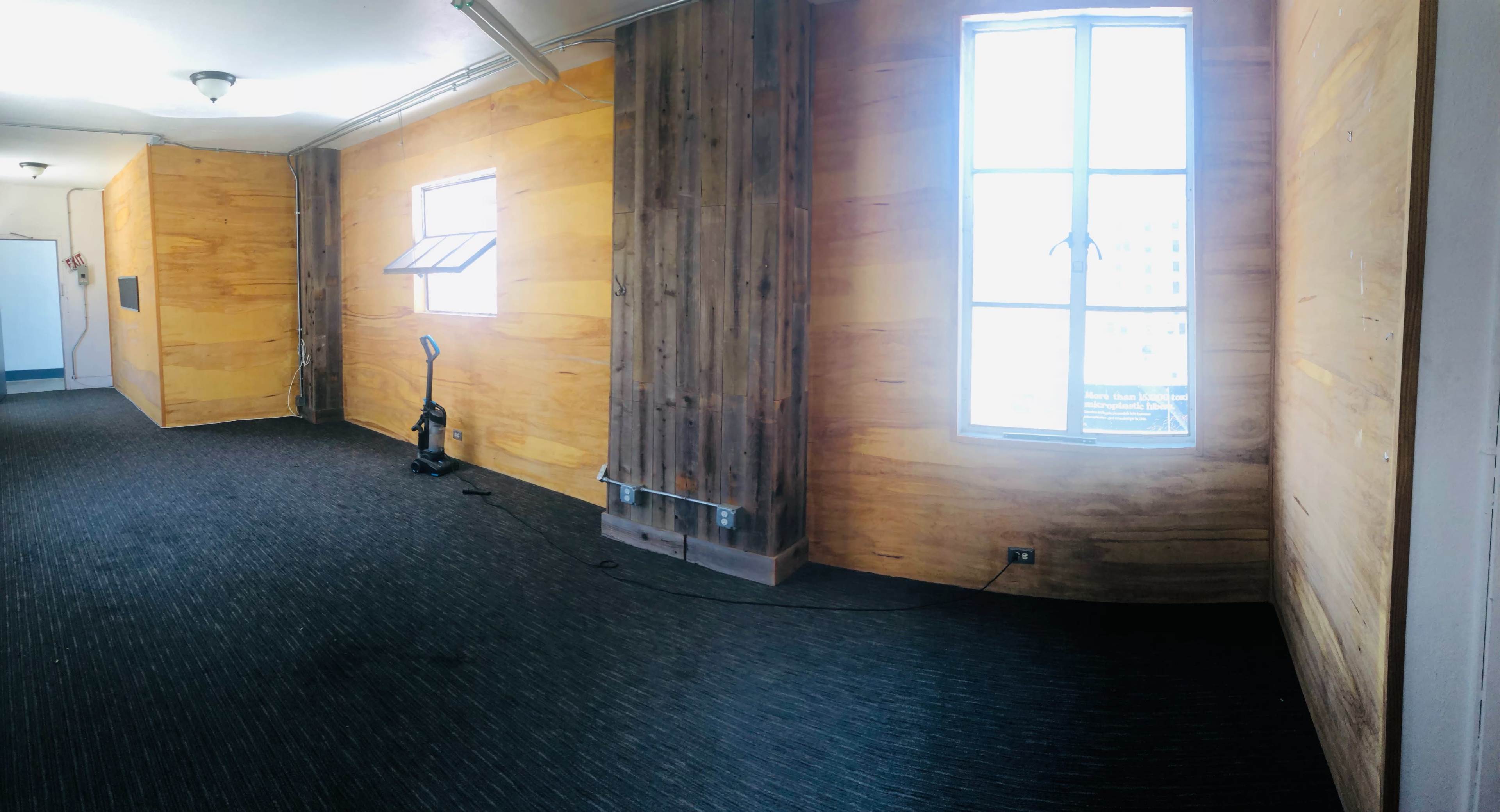 An empty room with wooden walls, a window, and a vacuum cleaner in the corner.