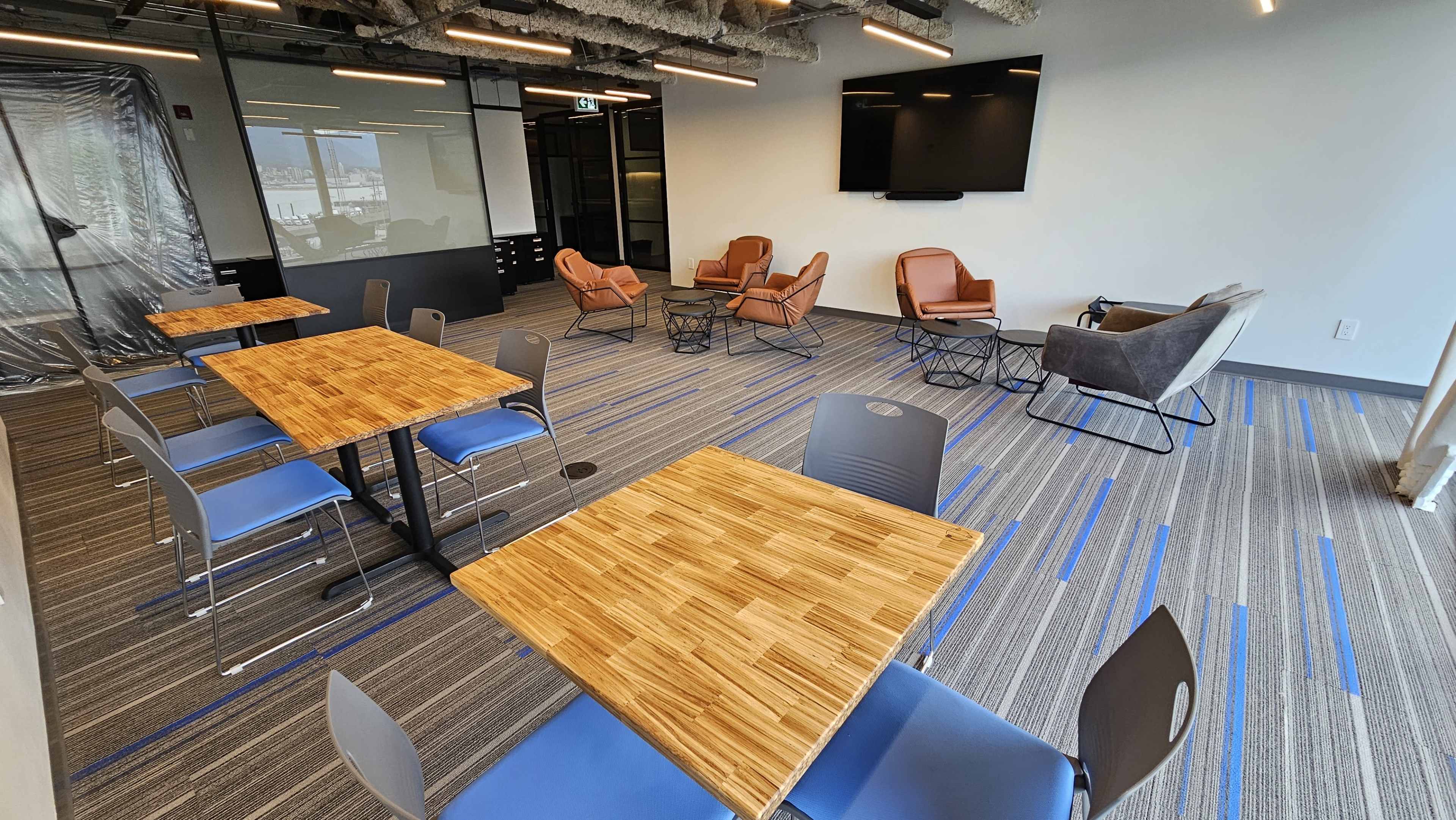 The image shows a modern office space with several wooden tables and chairs, a seating area with armchairs and a couch, and a large screen on the wall.