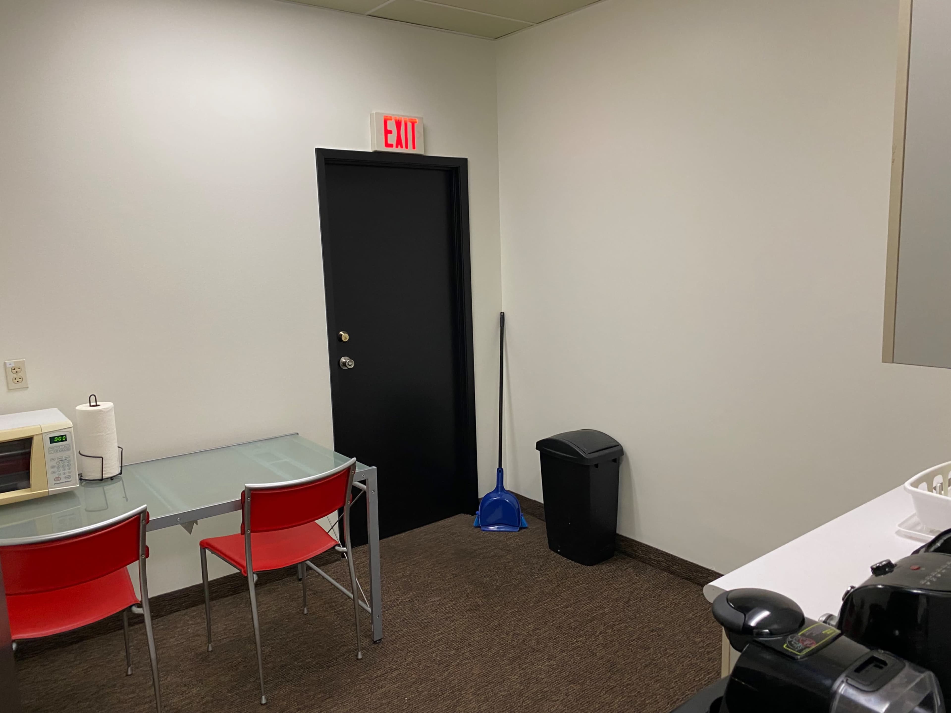 A small room with a table and red chairs, a door, a broom, and a trash can.