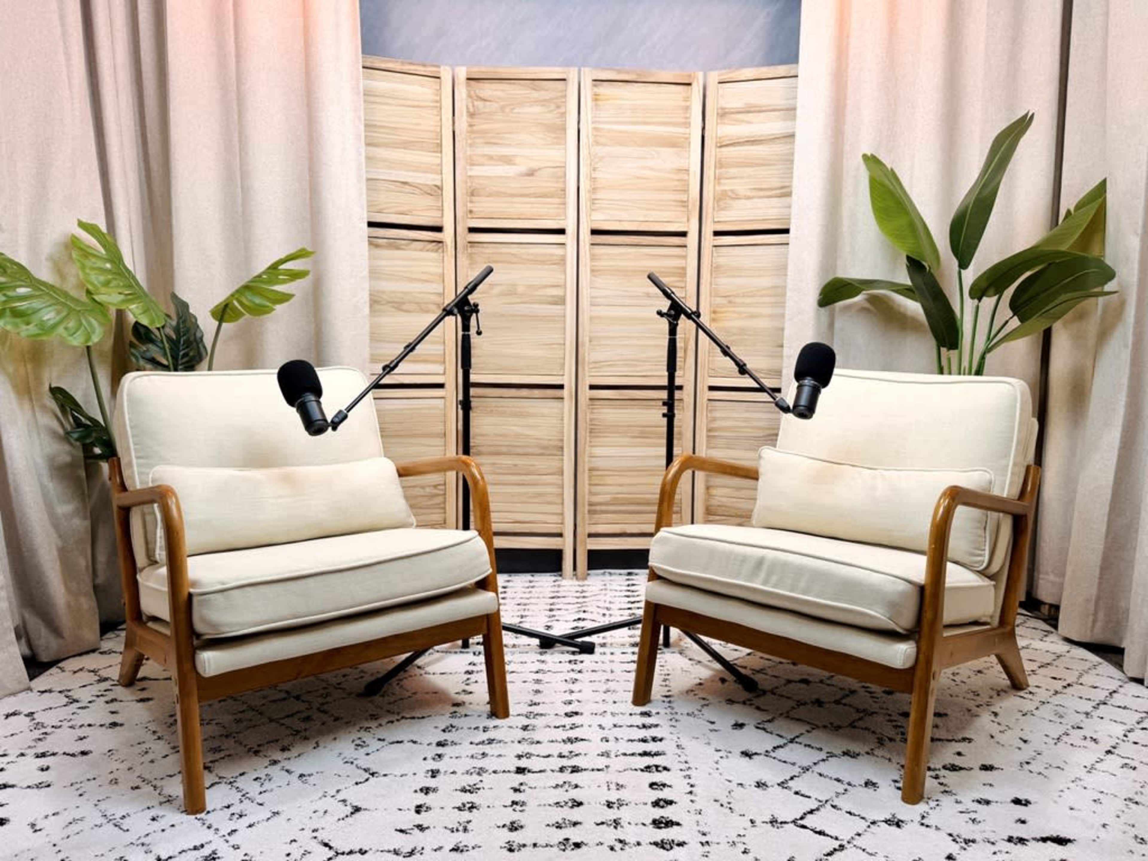 The image shows two light-colored chairs with microphones positioned in front of a wooden panel and surrounded by potted plants.