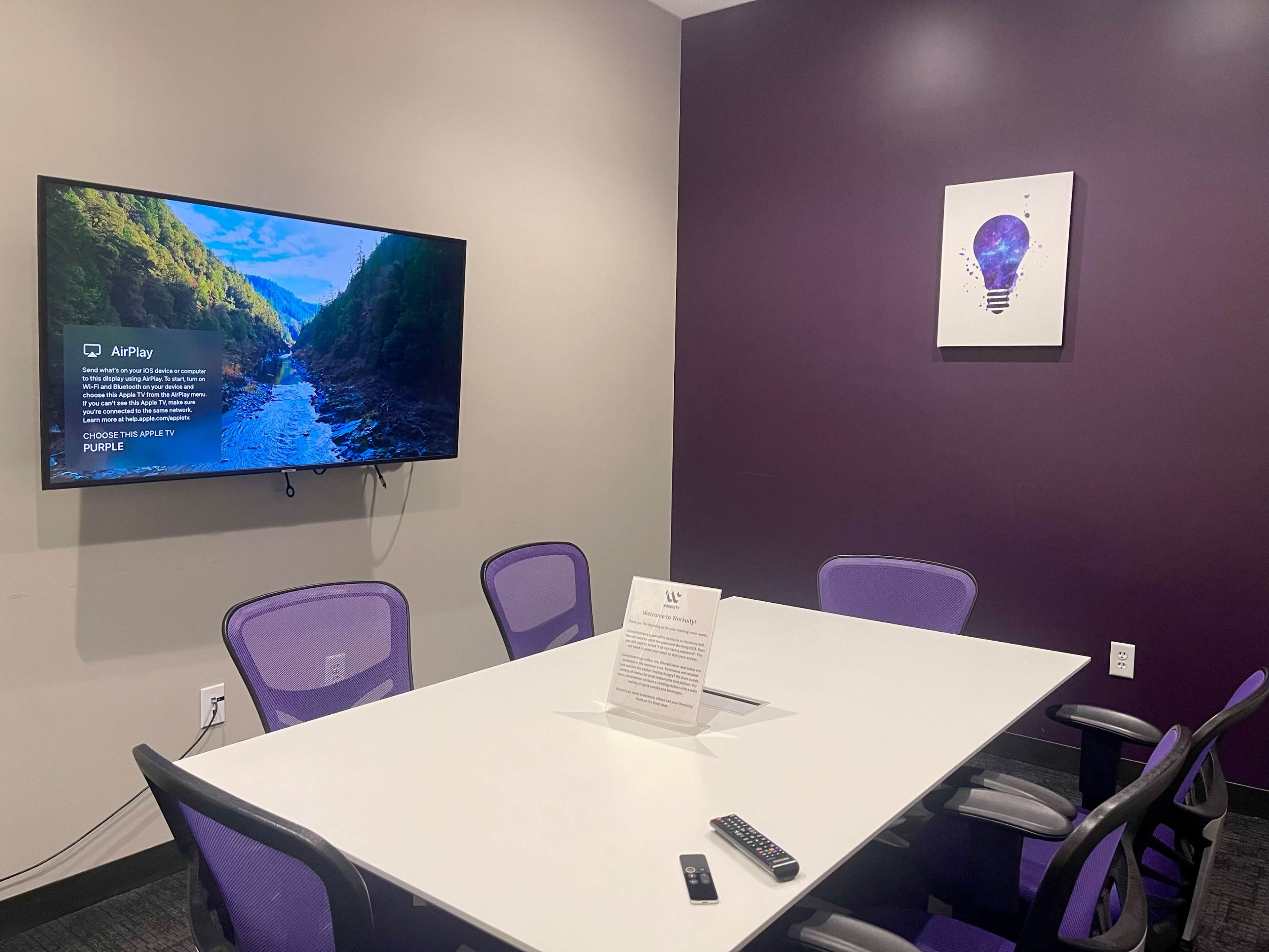 A conference room features a white table surrounded by purple chairs, a large TV displaying a nature scene, and a lightbulb artwork on the wall.