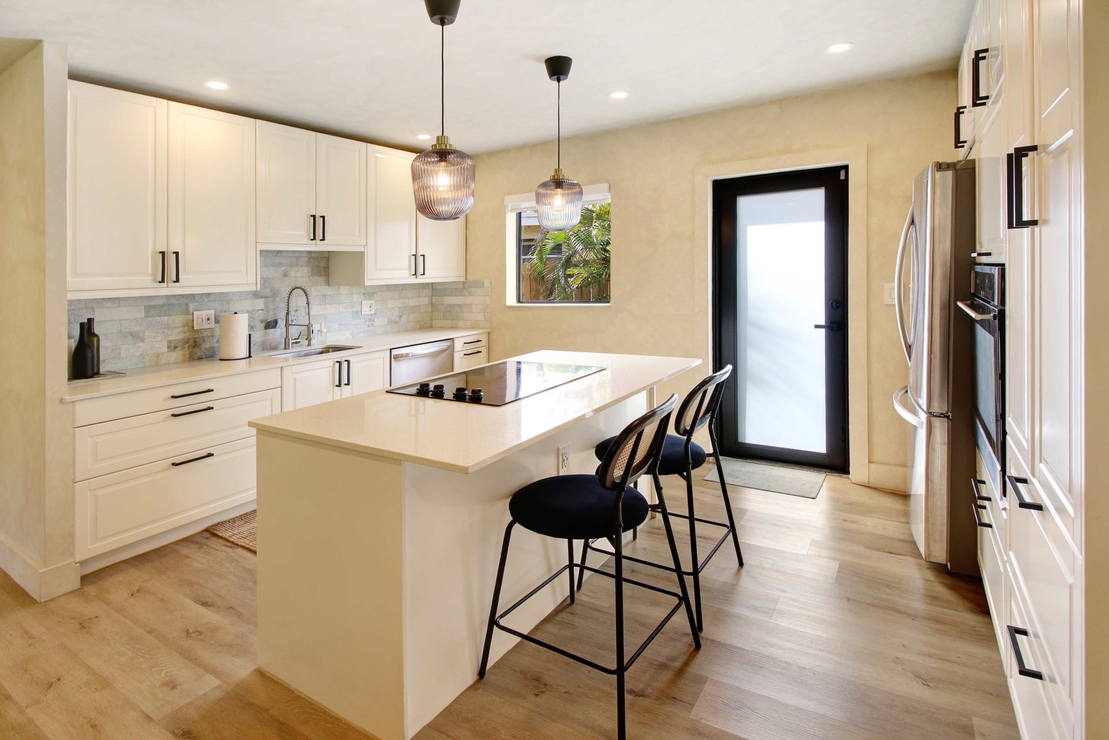 The image shows a modern kitchen with white cabinets, a central island with bar stools, and a sliding door leading to an outdoor area.