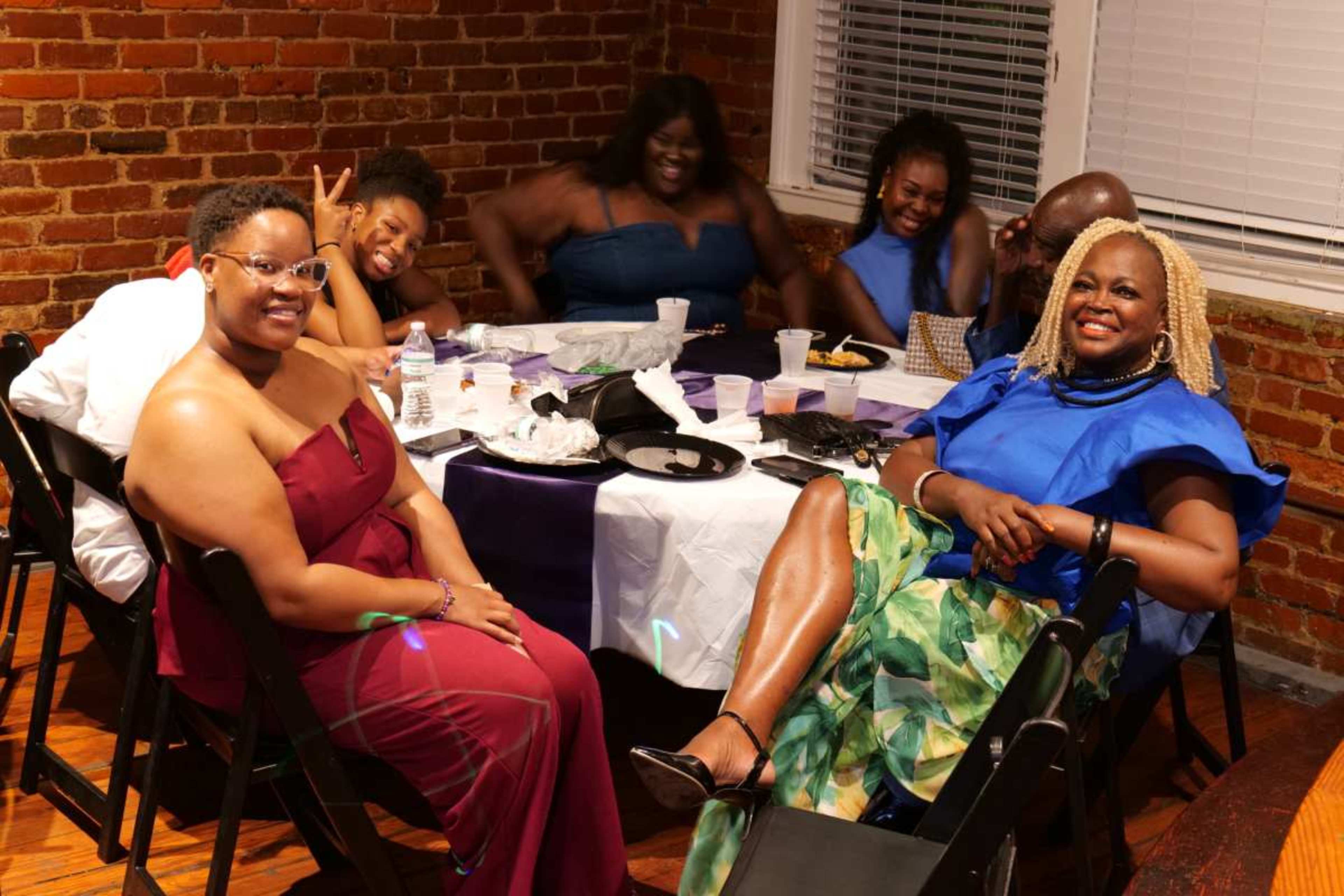 A group of six people are sitting around a dining table, smiling and enjoying a meal in a brick-walled room.