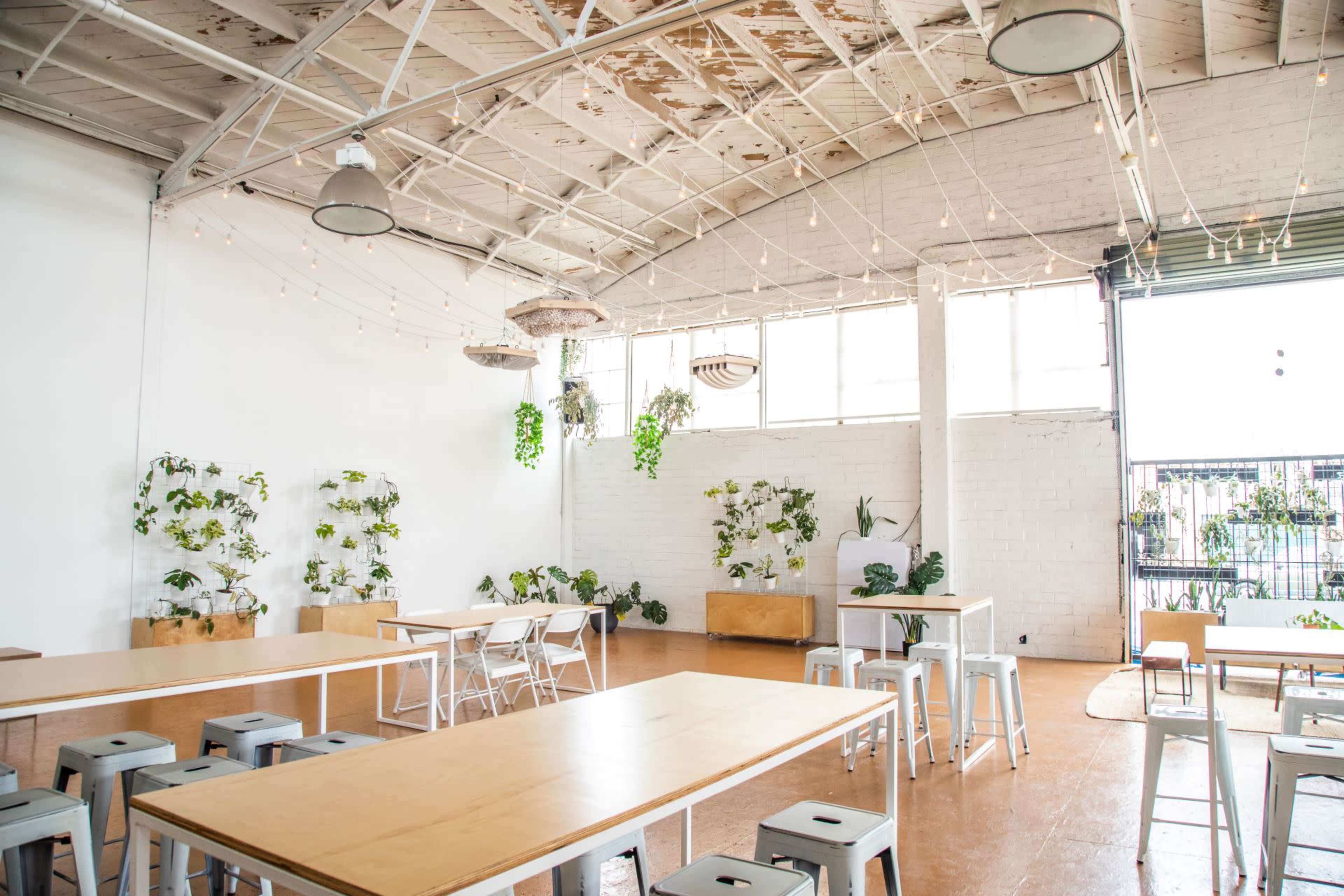 A spacious room features long wooden tables surrounded by metal stools, with plants along the walls and overhead string lights.