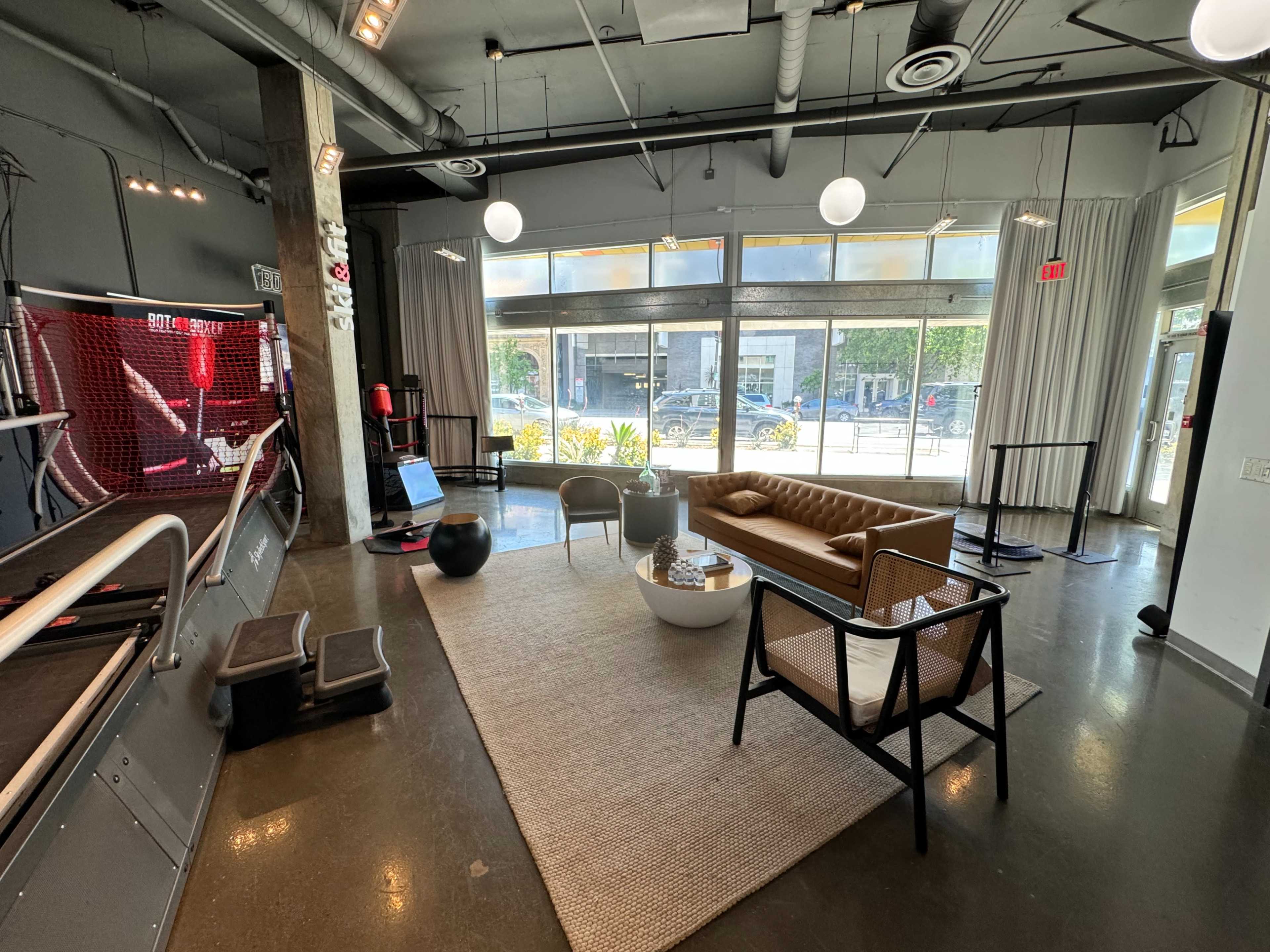 The image shows a modern interior space with a leather sofa, coffee table, and fitness equipment, featuring large windows that let in natural light.