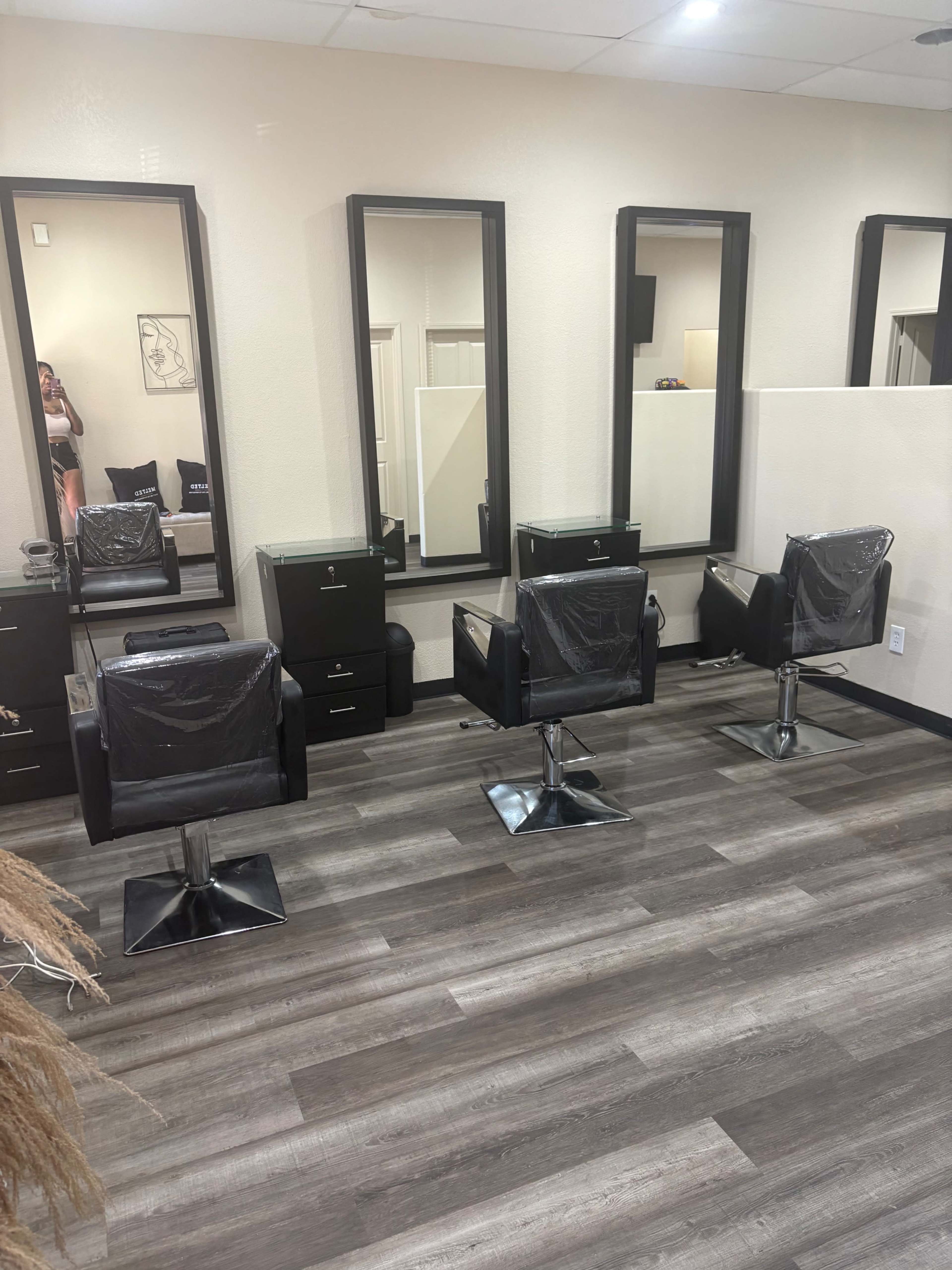 The image shows a hair salon interior with three black salon chairs positioned in front of tall mirrors and storage units.