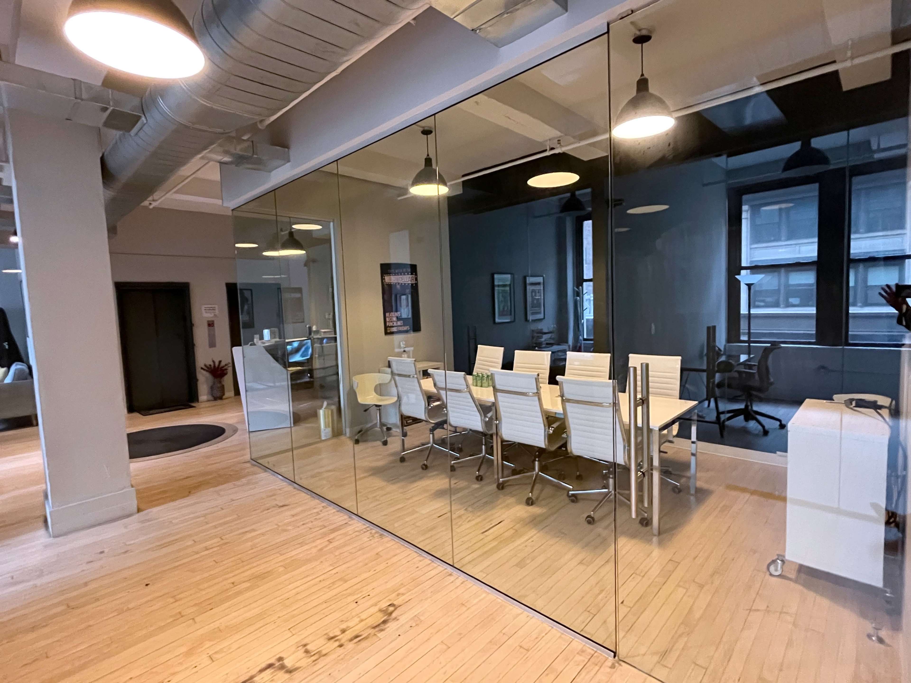 The image shows a modern conference room enclosed by glass walls, featuring a long table surrounded by white chairs and two additional office spaces visible in the background.
