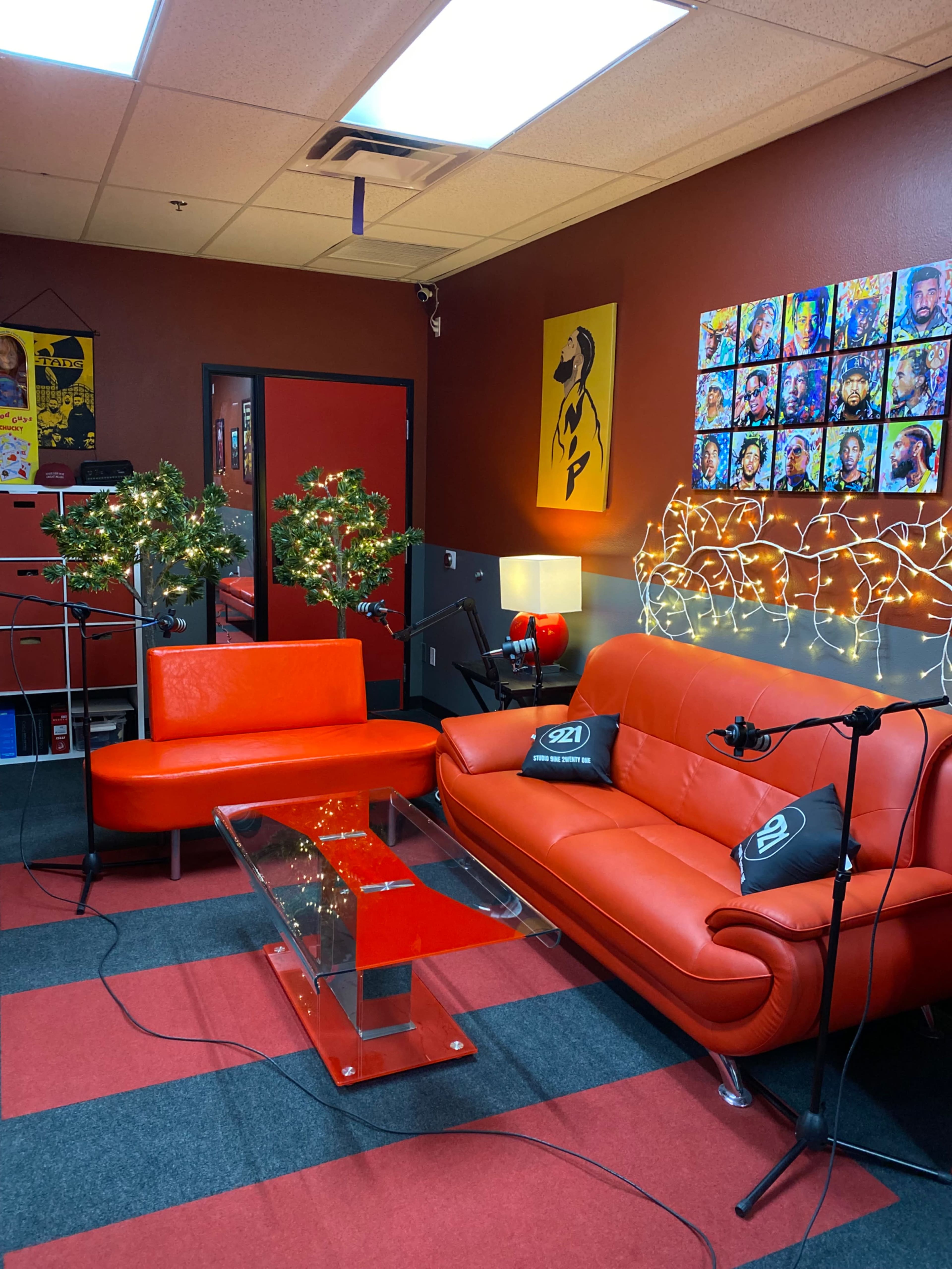 The image shows a recording studio with red walls, orange furniture, decorative lighting, and microphones set up for a podcast.