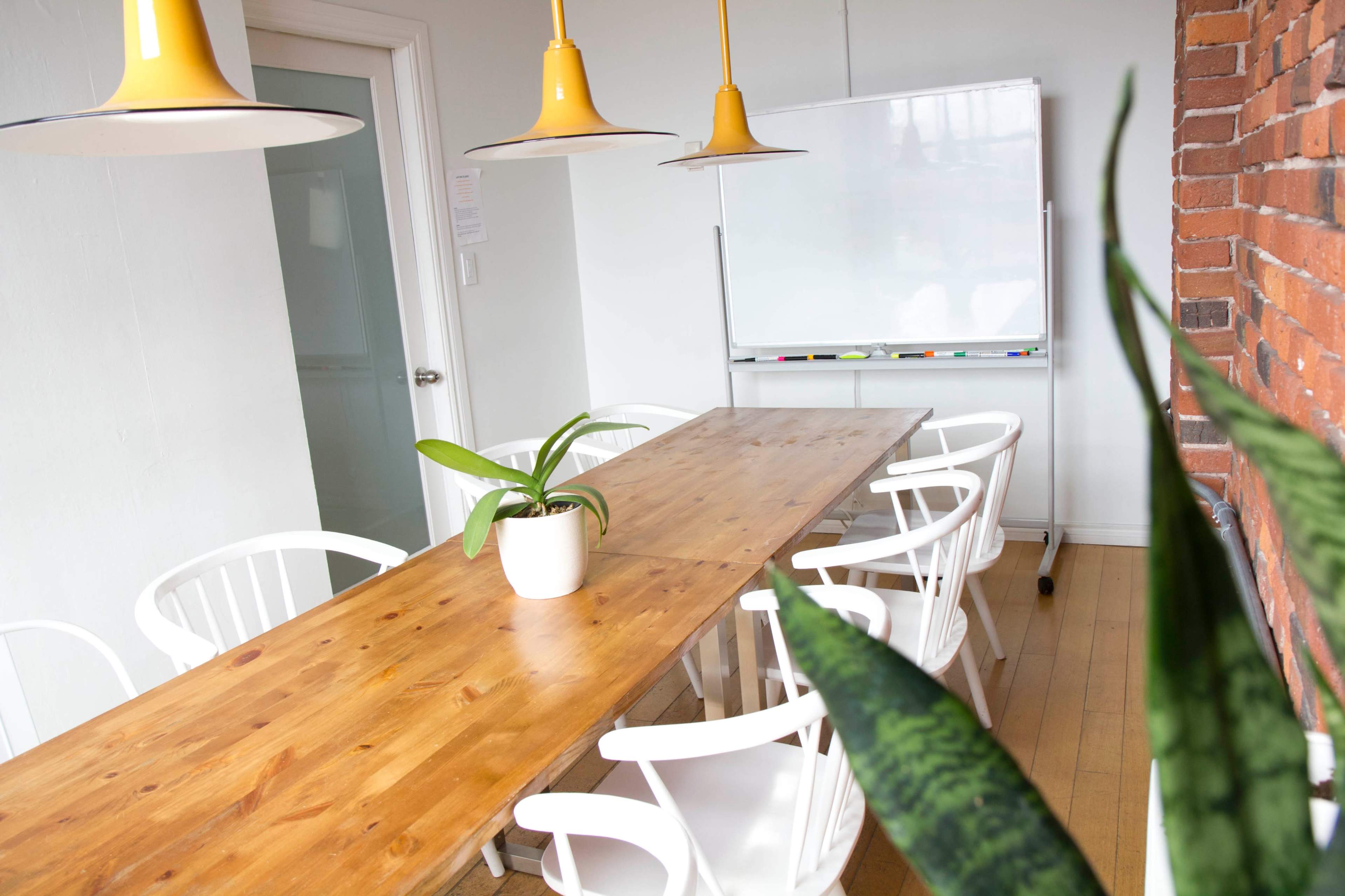 A simple conference room features a long wooden table surrounded by white chairs, with a potted plant and a whiteboard in the background.