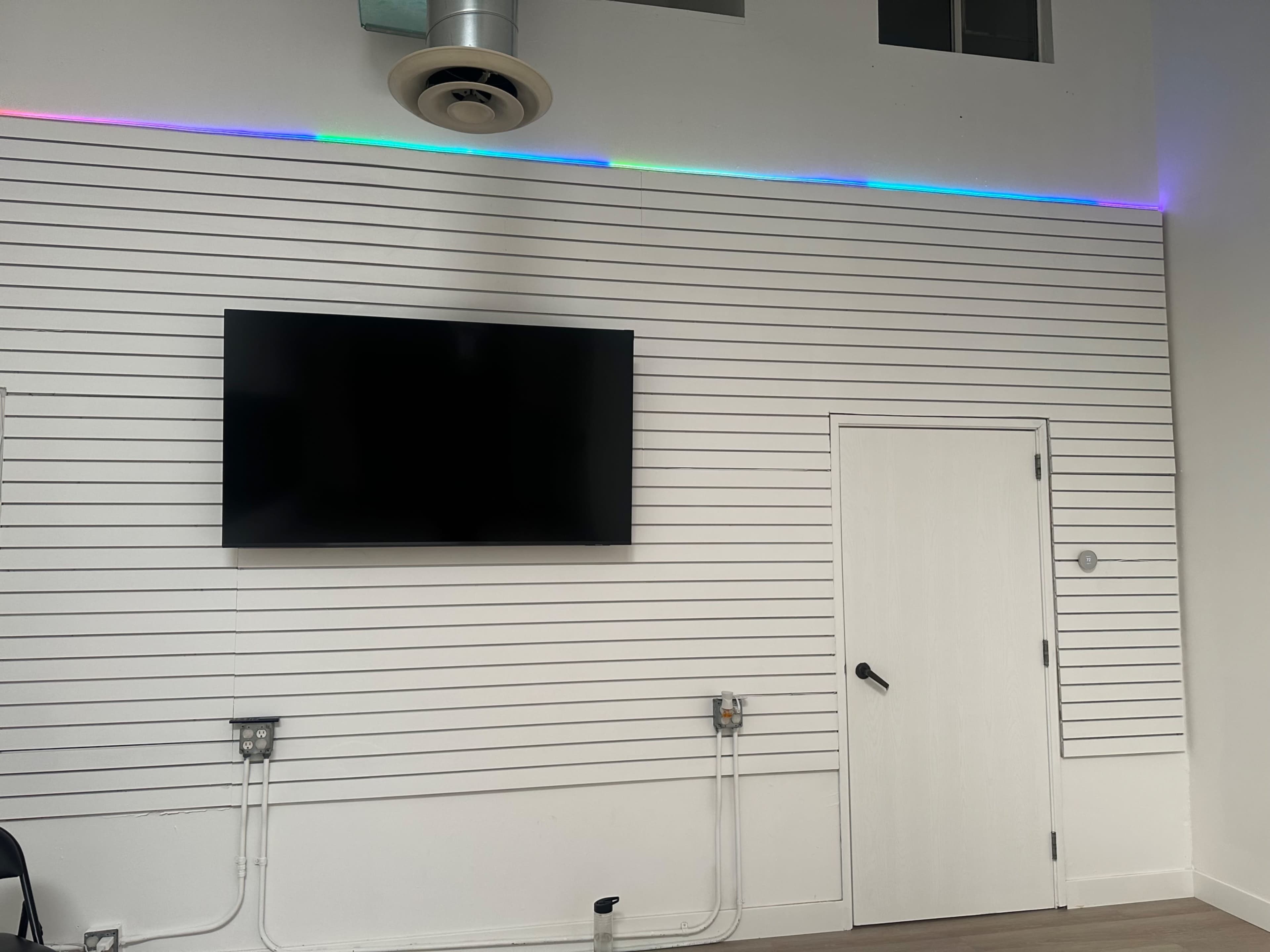 The image shows a white wall with horizontal paneling, a mounted flat-screen TV, a door, and a multicolored LED light strip near the top of the wall.