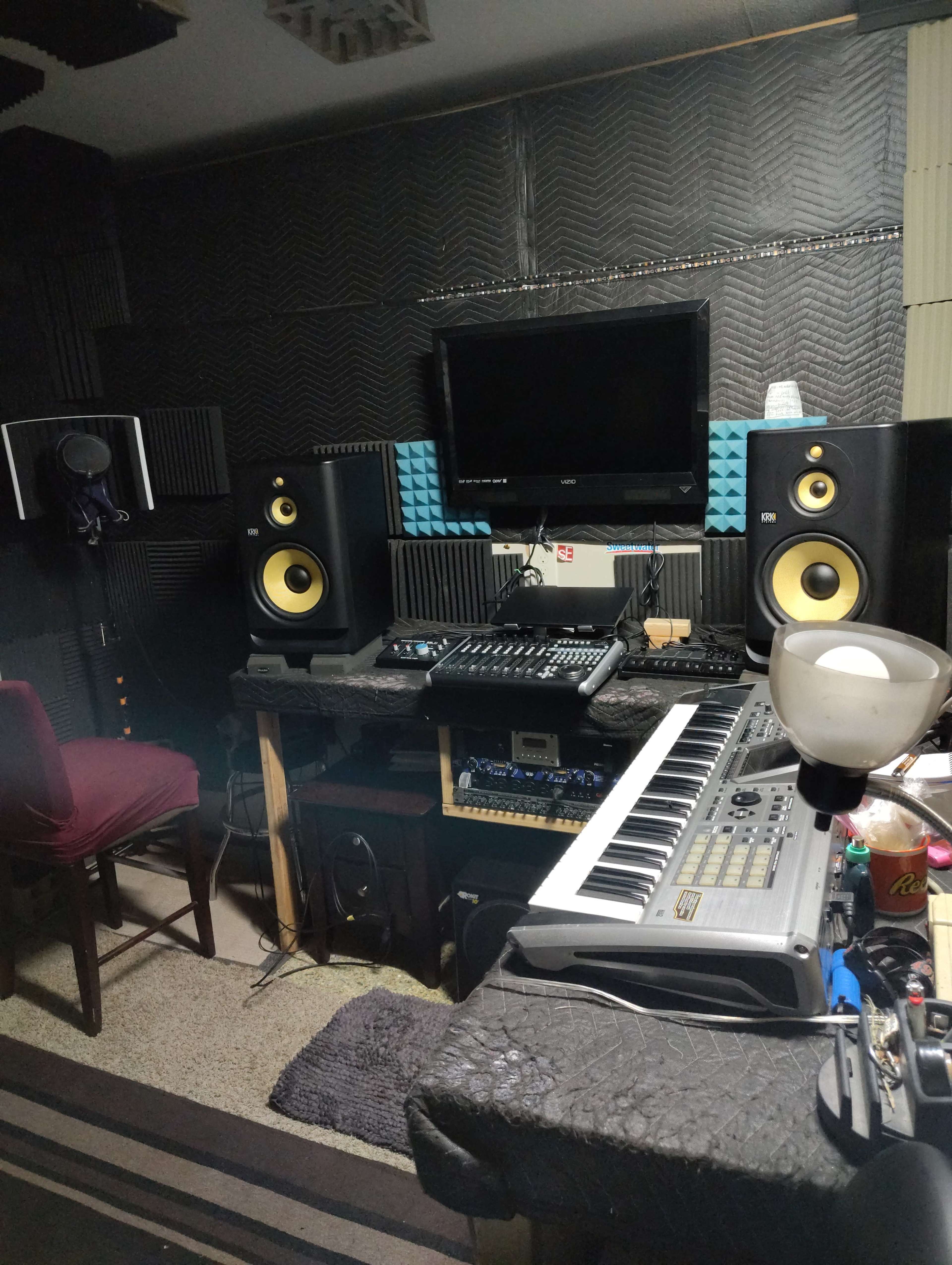 The image shows a music production studio with speakers, a mixing console, a keyboard, and soundproofing on the walls.