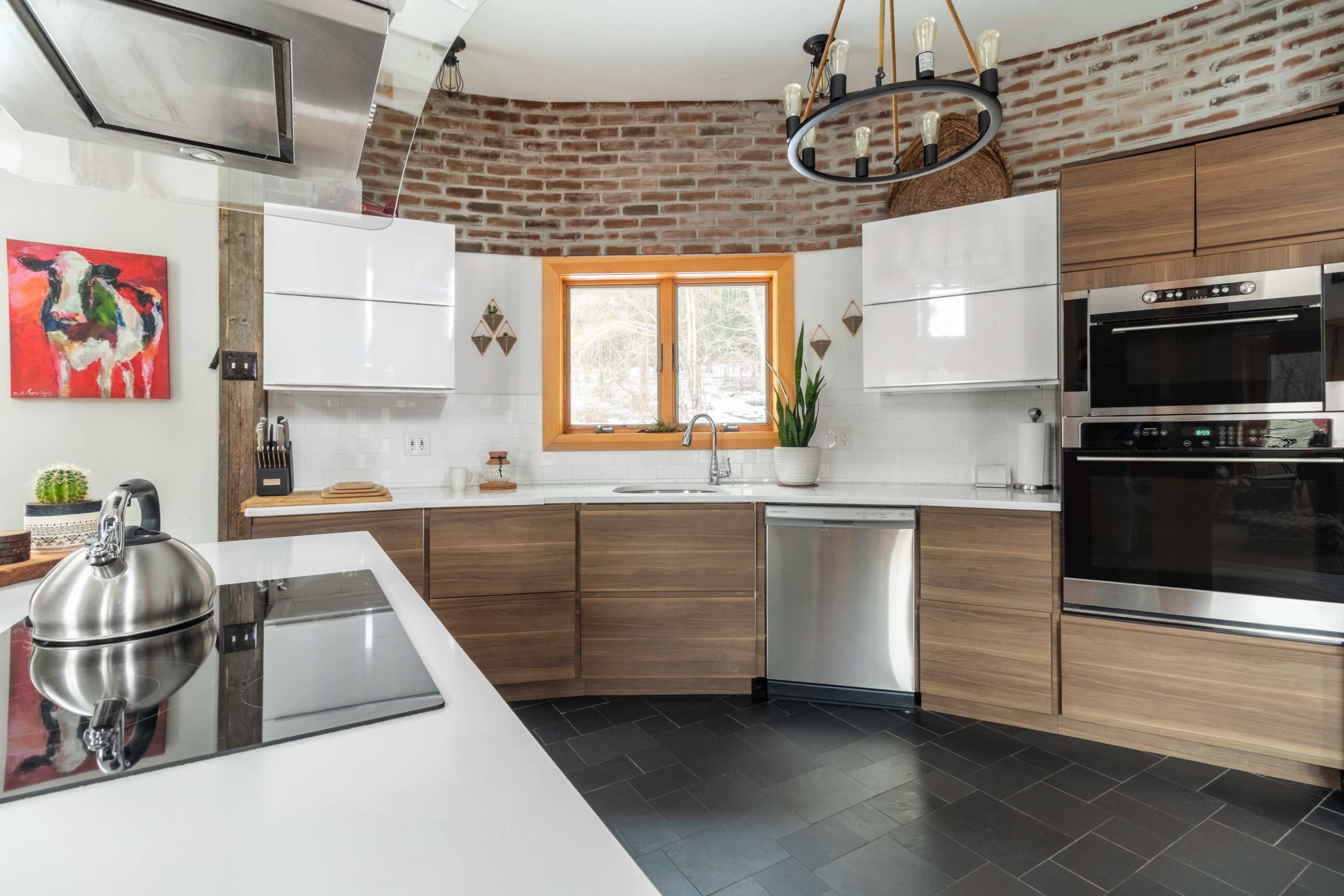 A modern kitchen features a circular layout with wooden cabinets, a stainless steel refrigerator, and a window overlooking greenery.