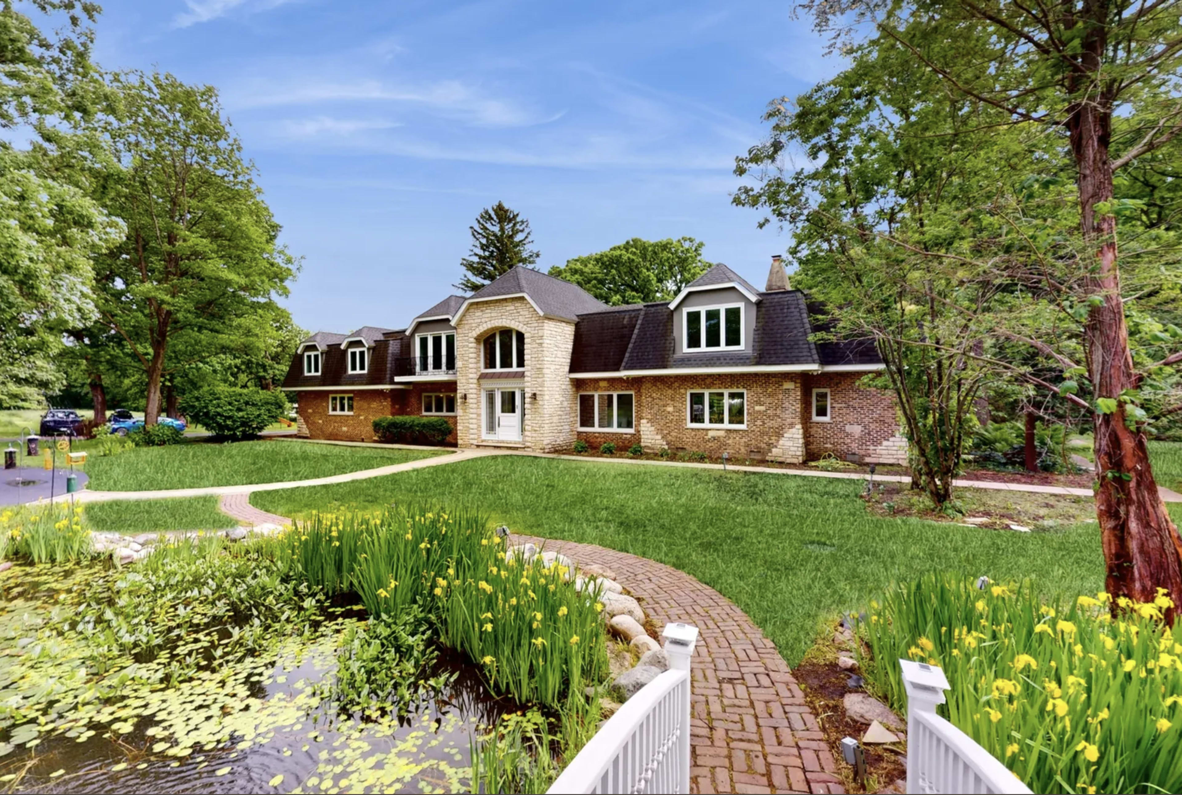 The image shows a large stone house with multiple windows, surrounded by green lawn and trees, alongside a pond with yellow flowers.